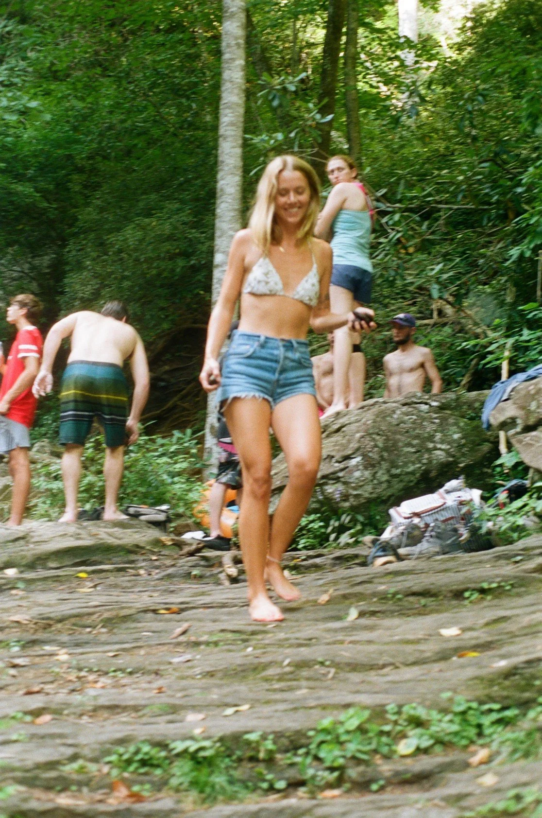 A group of people enjoying a day outdoors in a forest, with some standing on large rocks and others walking barefoot on a wooden trail. The central figure is a woman in a bikini top and denim shorts, smiling and holding a phone.