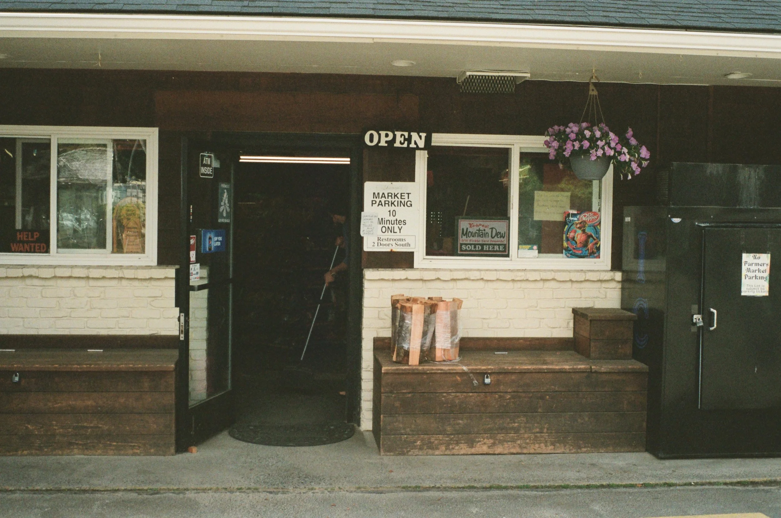 Front view of a small grocery store with signs indicating it is open, with hanging flower basket and various signs in the window, including 'Mountain Dew' and 'Help Wanted'