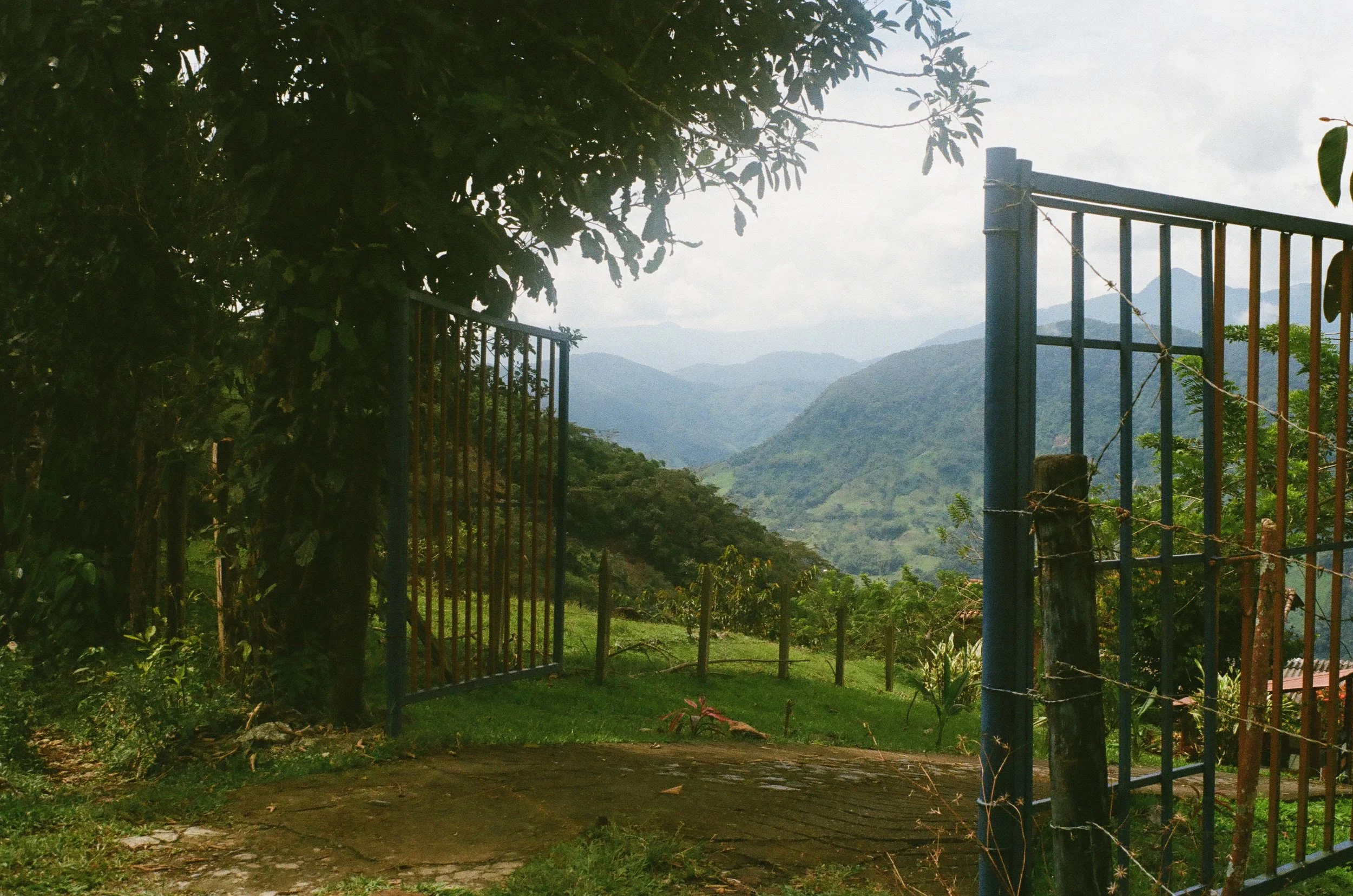 Open blue metal gate leading to a lush green mountain valley with rolling hills and cloudy sky in the background.