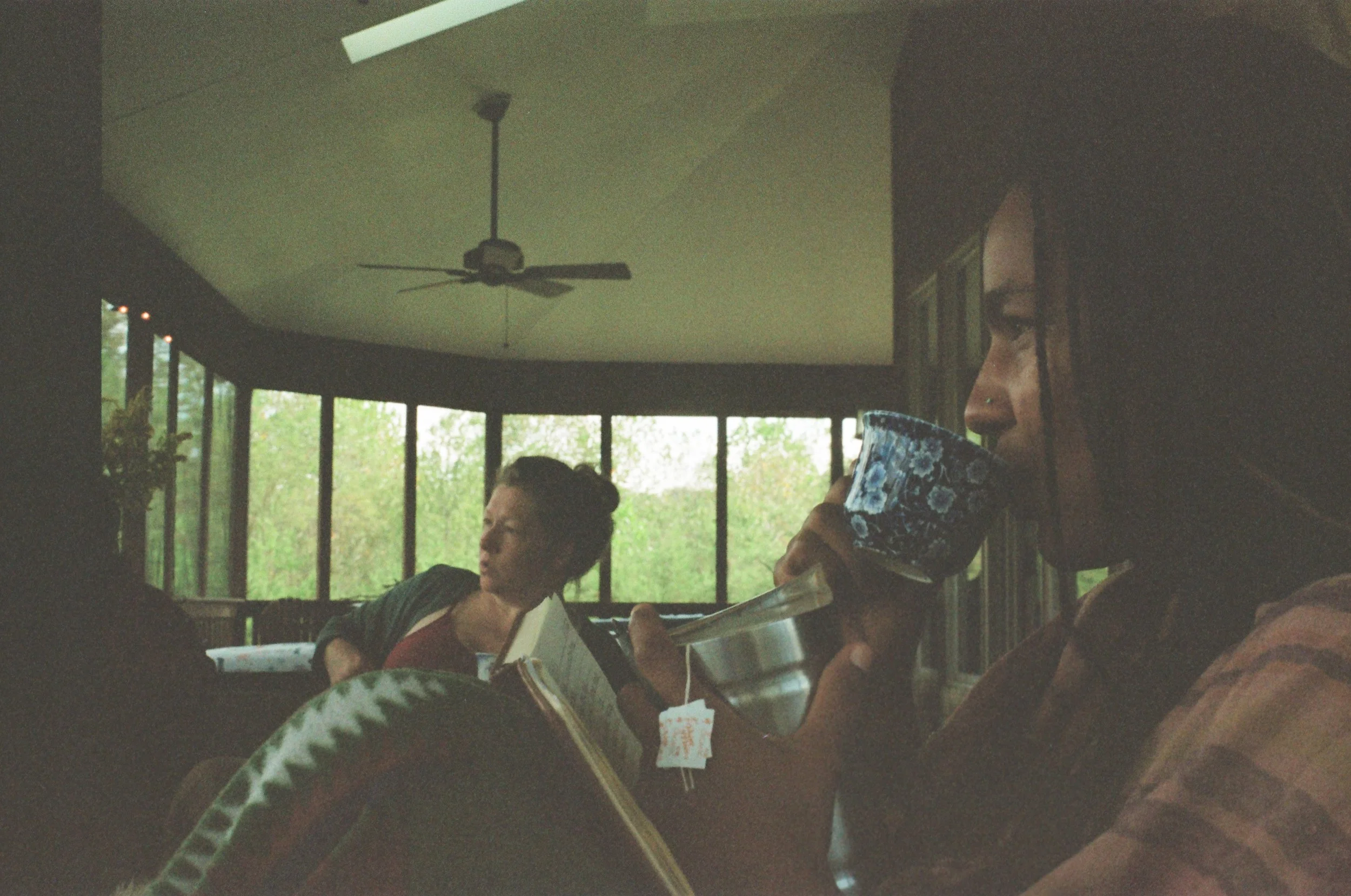 A woman with dark hair and a nose piercing drinks from a mug while sitting at a table, with another woman in the background reading a book, in a room with large windows and a ceiling fan.
