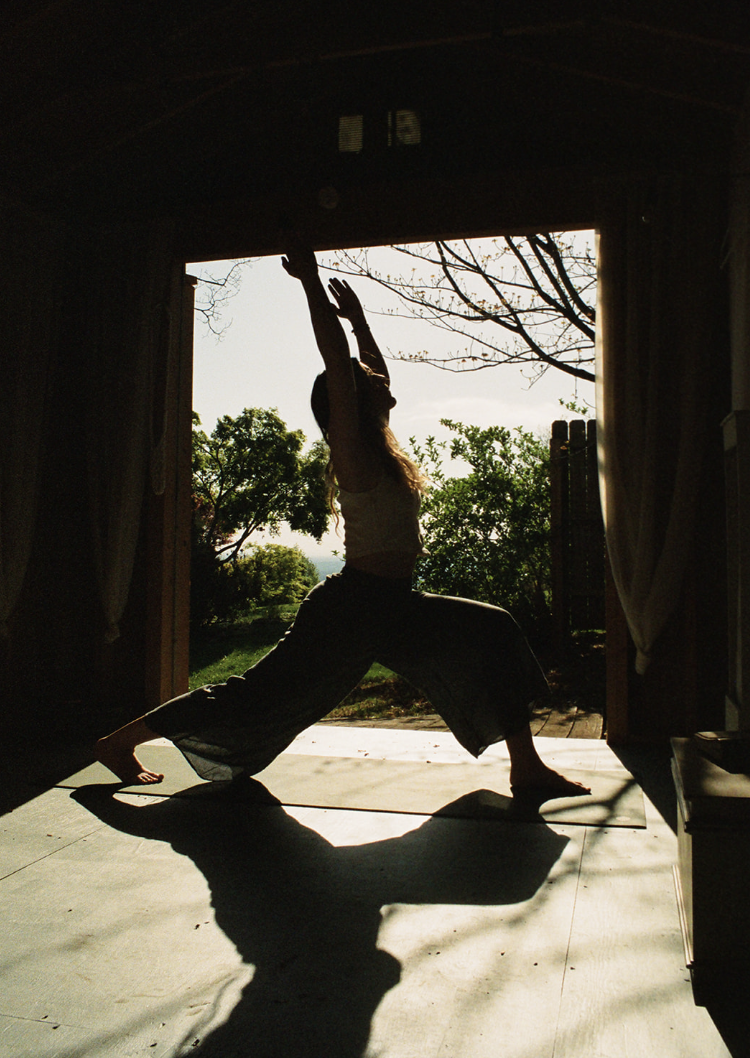 A person practicing yoga in a wide stance with arms raised overhead, silhouetted against a bright open doorway with outdoor trees and sky visible.