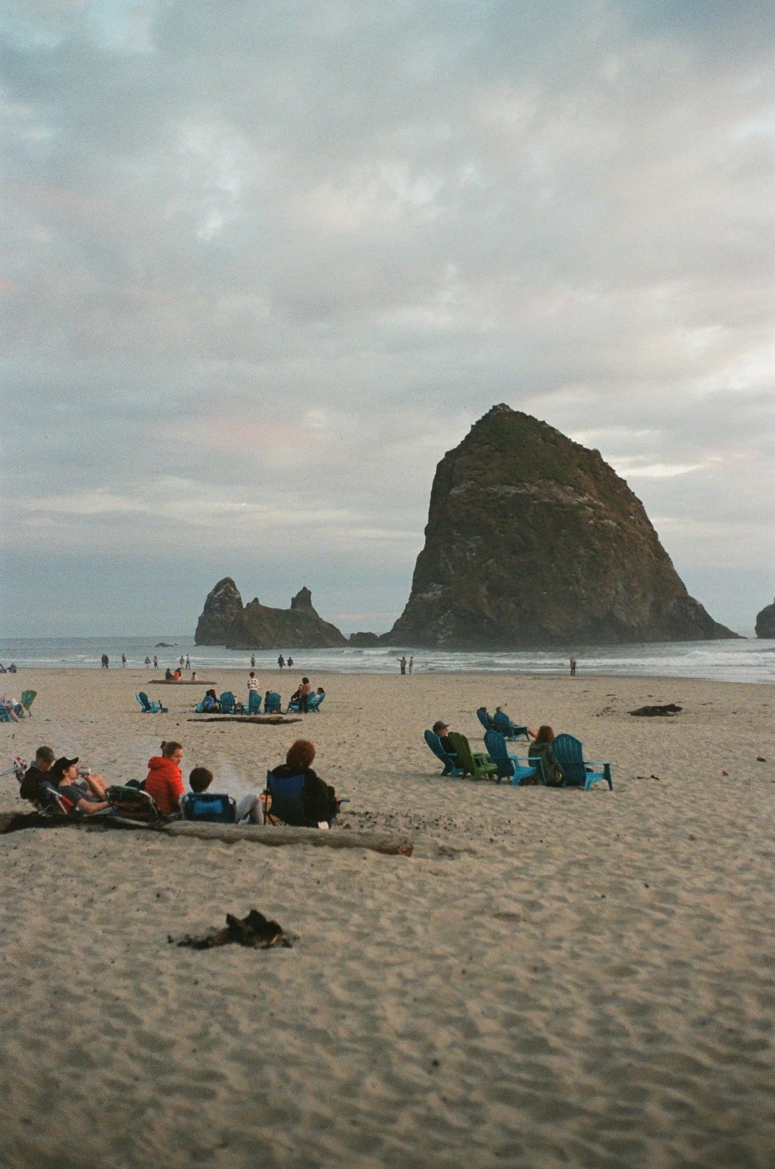 People sitting on beach chairs and blankets on a sandy beach, with large rock formations emerging from the ocean in the background, under a cloudy sky.