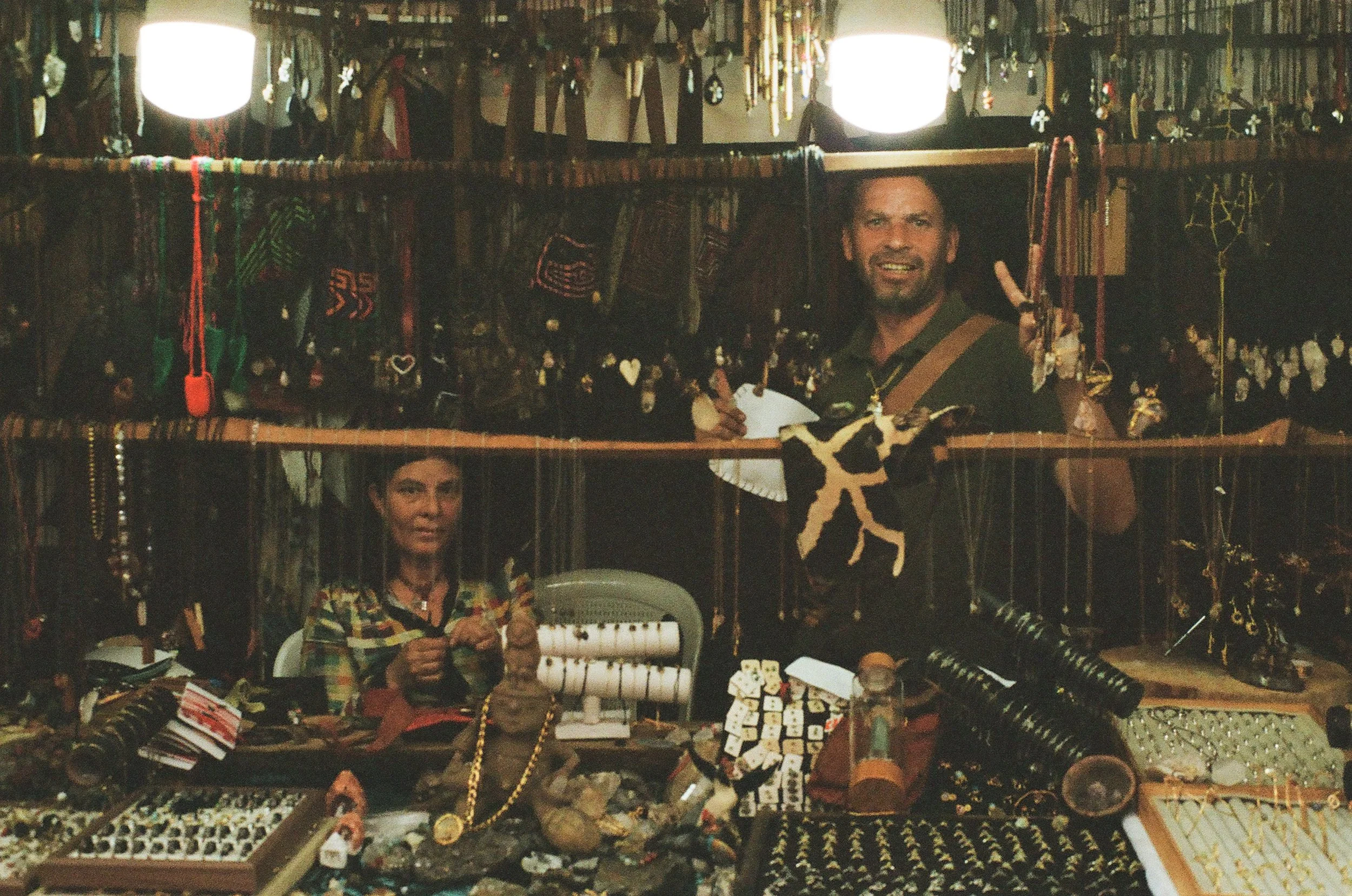 A man and woman shopping at a jewelry stall in a market. The stall displays various jewelry items, including necklaces, bracelets, and charms, with some crafted from beads and stones. The man is making a peace sign with his fingers. The scene is insi