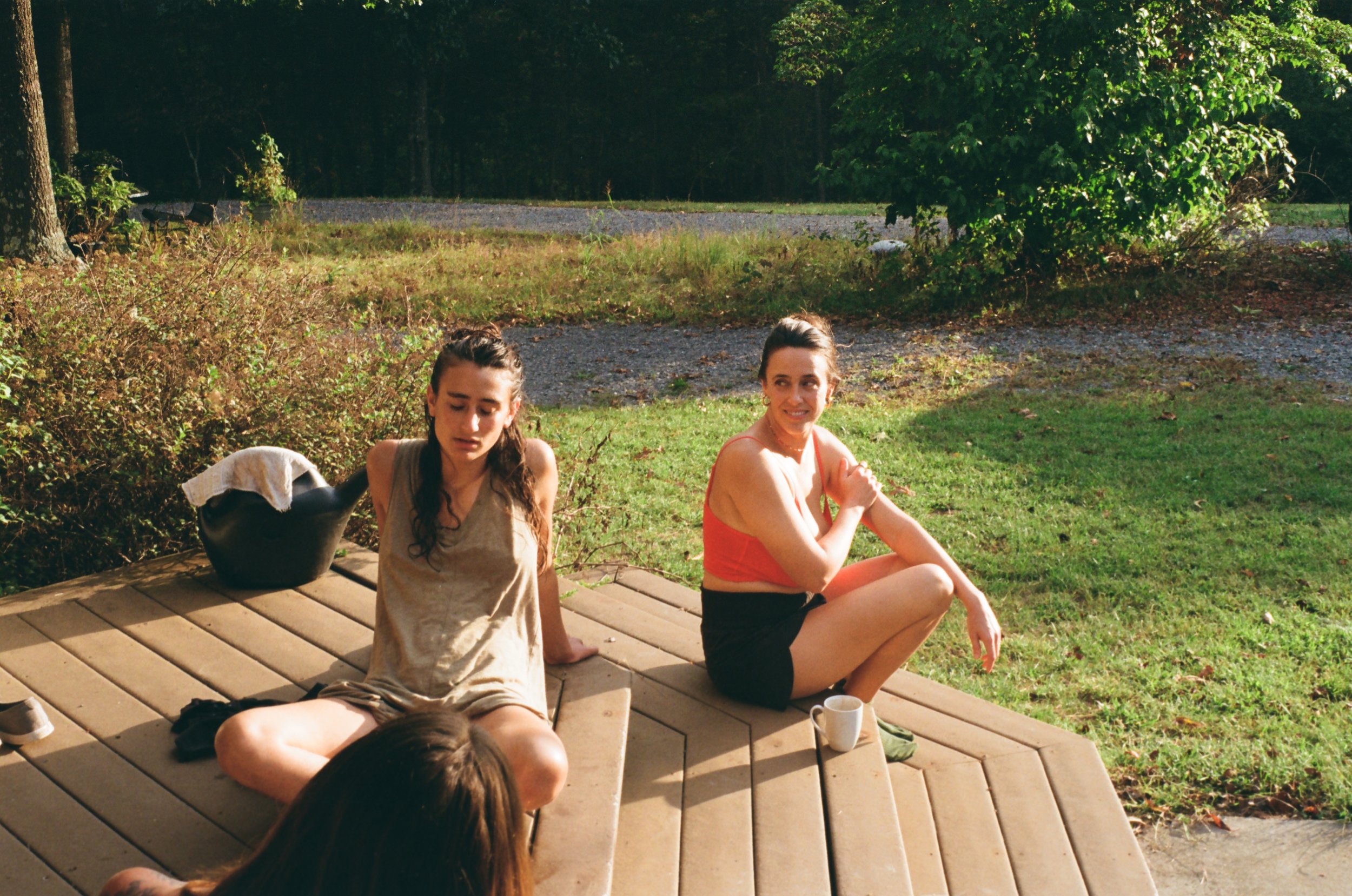 Two women sitting on a wooden deck outdoors, one with long dark hair in a beige sleeveless top and the other with dark hair in a red top and black shorts, with a background of green trees and grass.