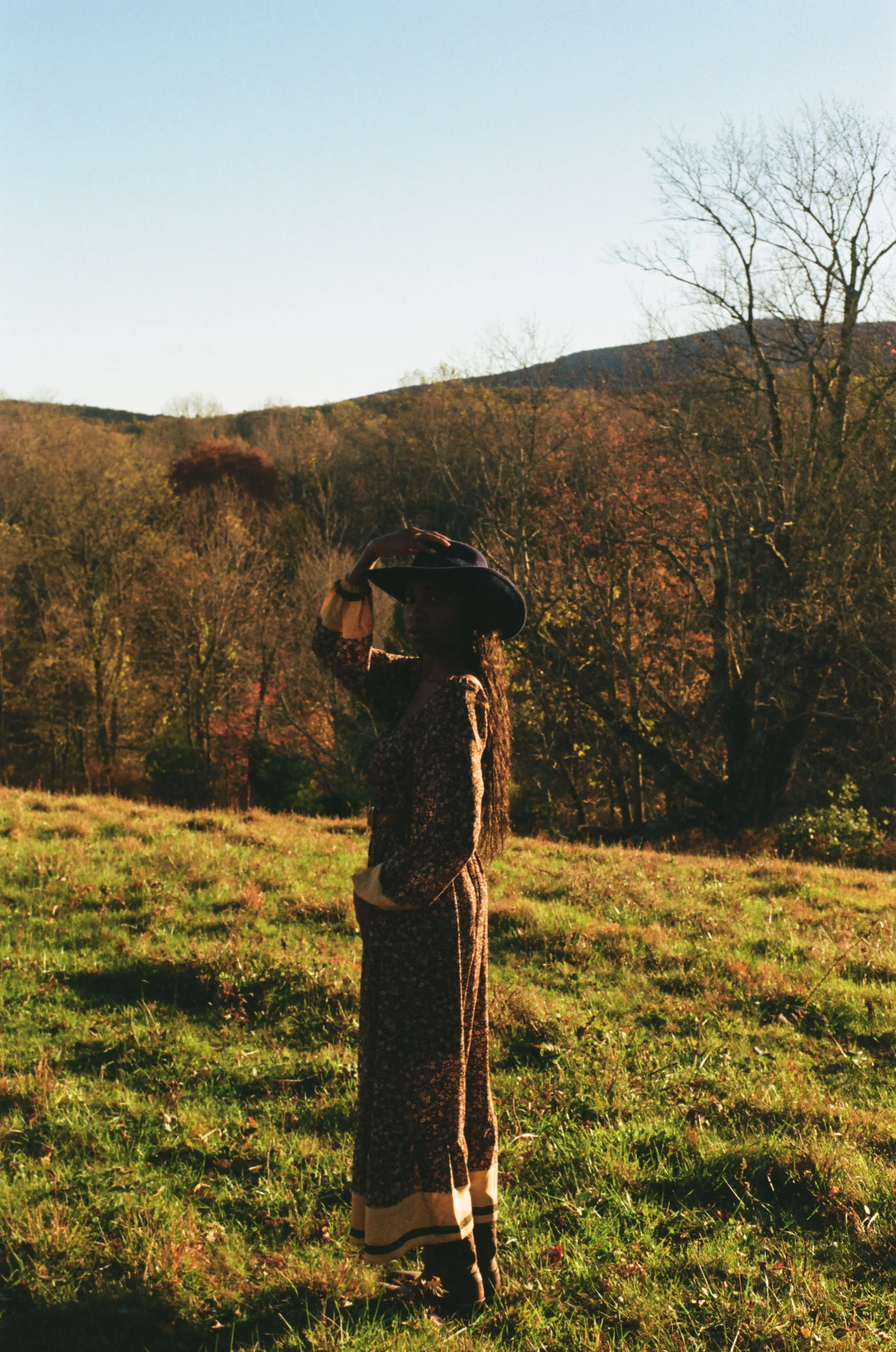 A woman wearing a floral dress and a wide-brimmed hat standing outdoors on a grassy field, with a backdrop of leafless trees and hills under a clear sky.
