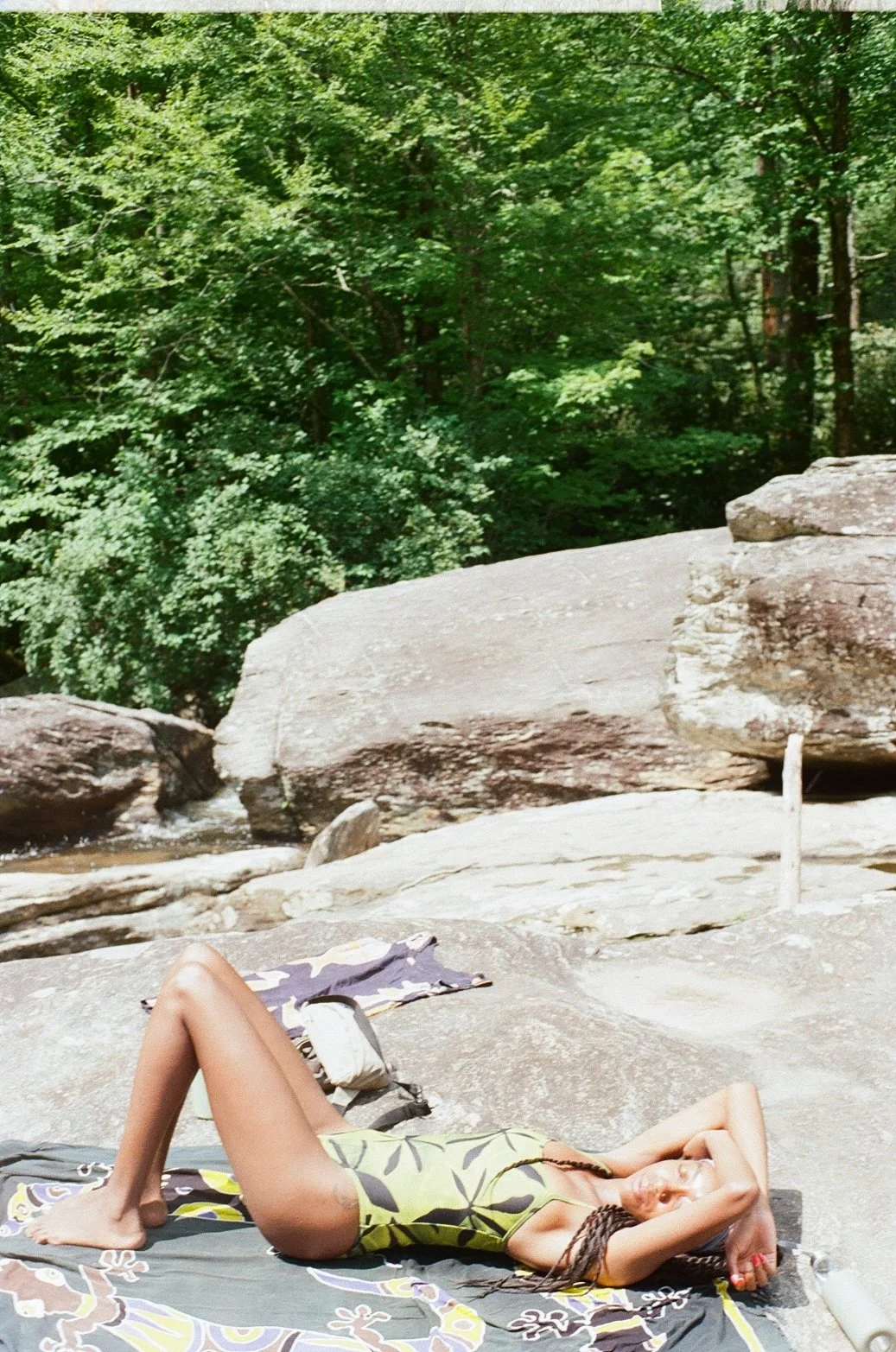 A woman with braided hair lying on a towel on a large, flat rock in a forested area, with lush green trees in the background.