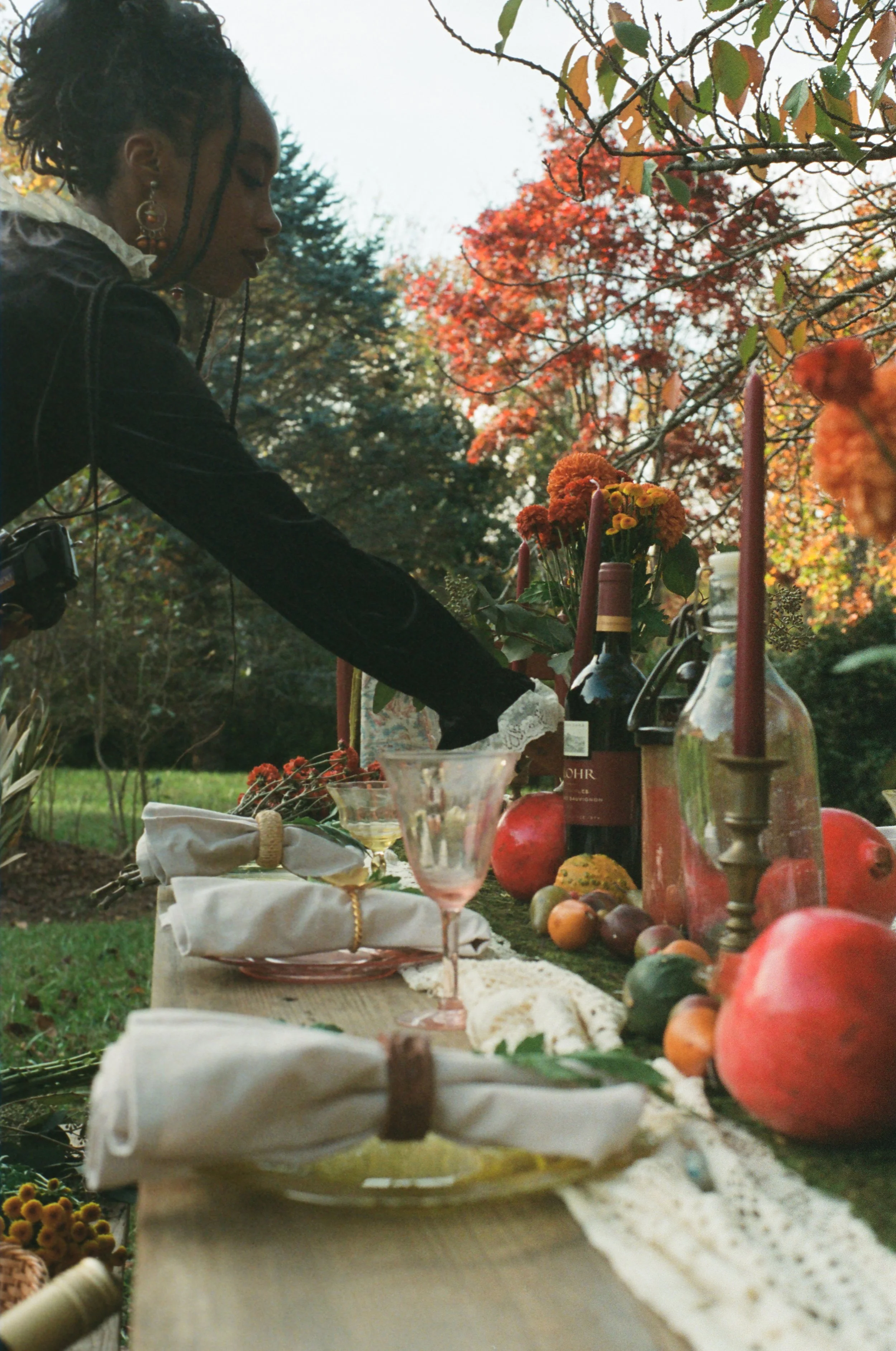 A woman setting a table outdoors with fall decor, including flowers, pomegranates, and wine bottles, in a garden with autumn foliage.