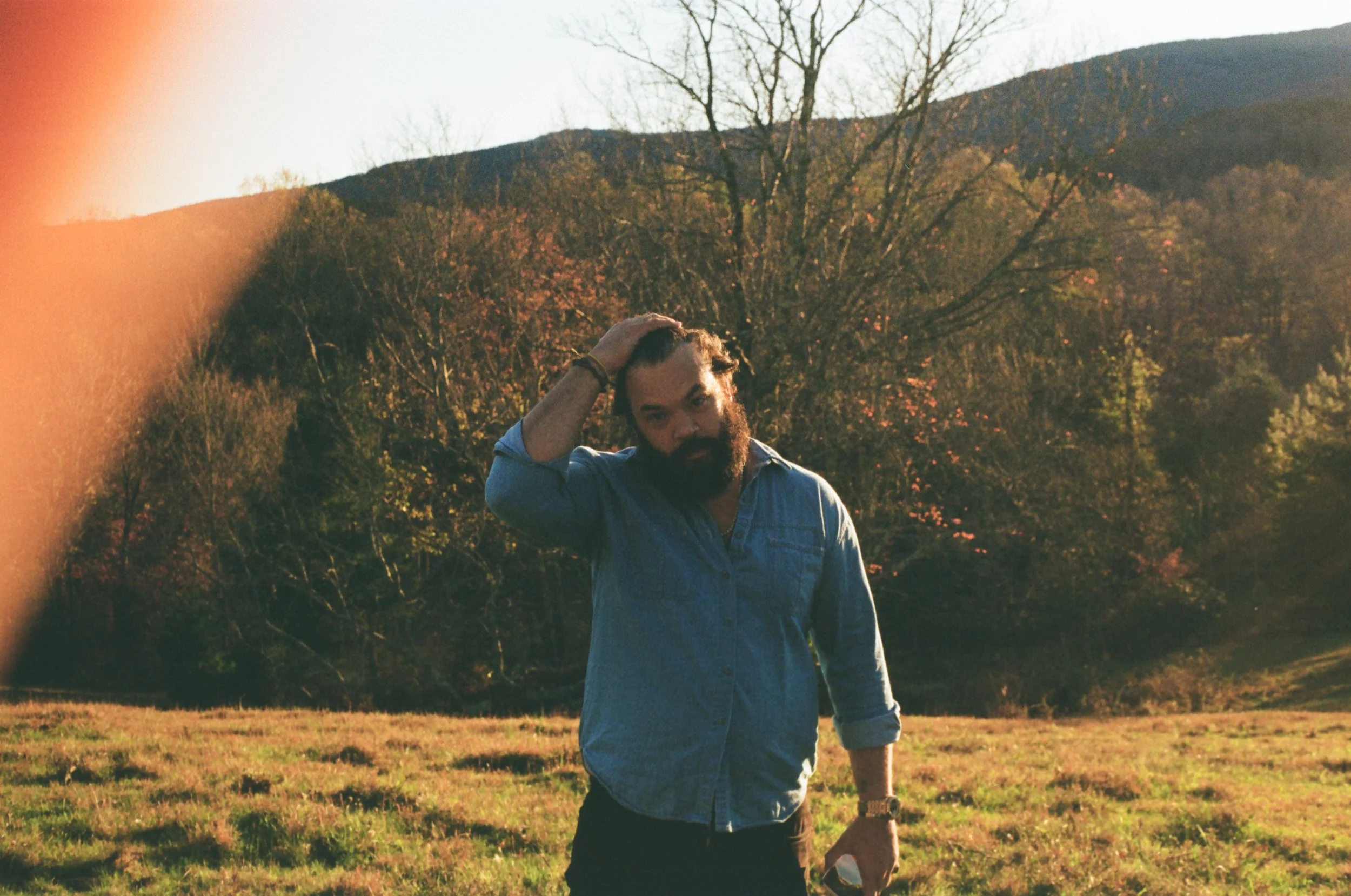 A man with a beard, wearing a denim shirt, is standing outdoors in a grassy field with trees and mountains in the background. He is touching his head with one hand and holding sunglasses in the other. The scene appears to be during the late afternoon