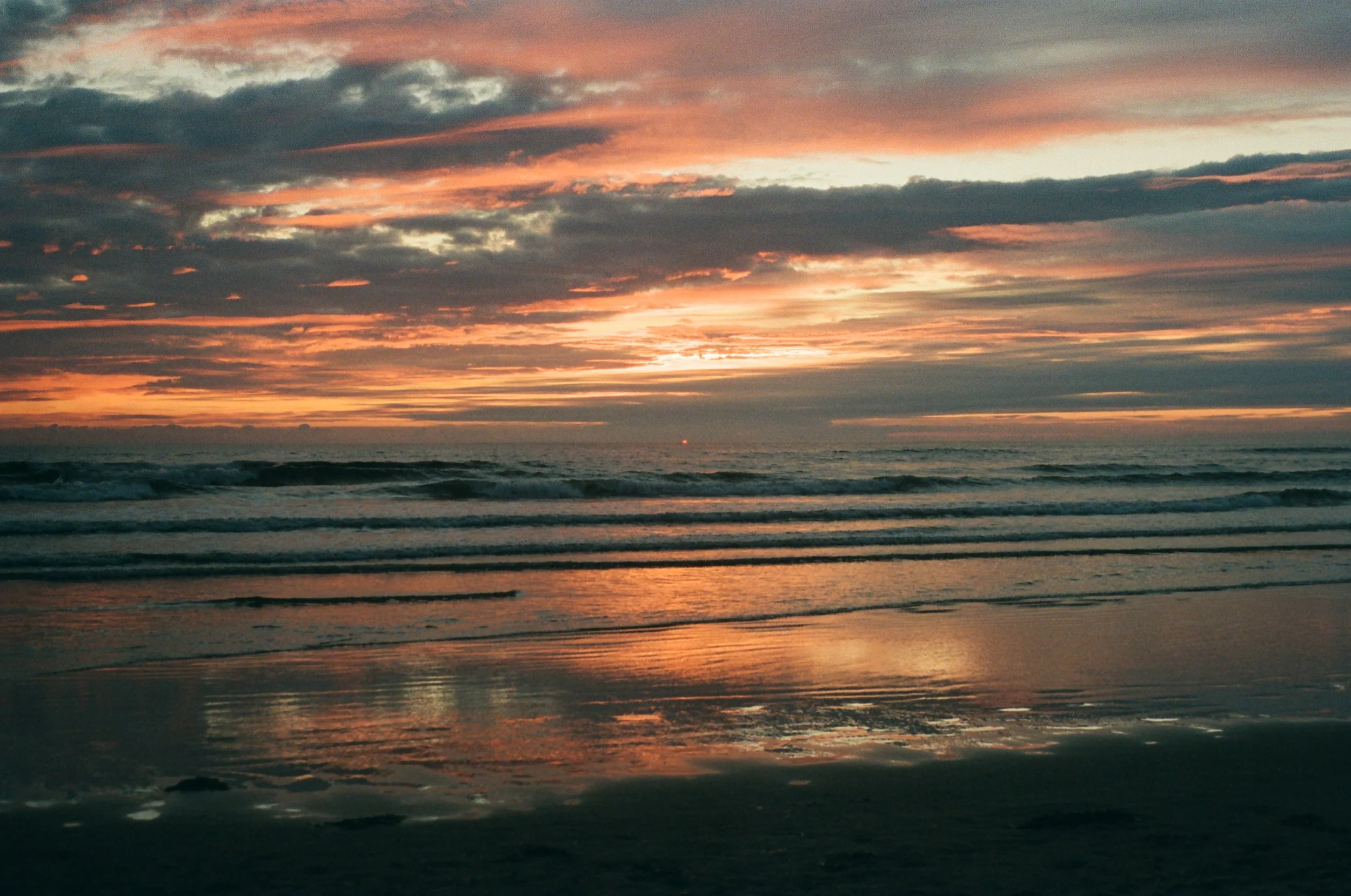 Sunset over the ocean with clouds and reflected colors on the wet sandy beach.