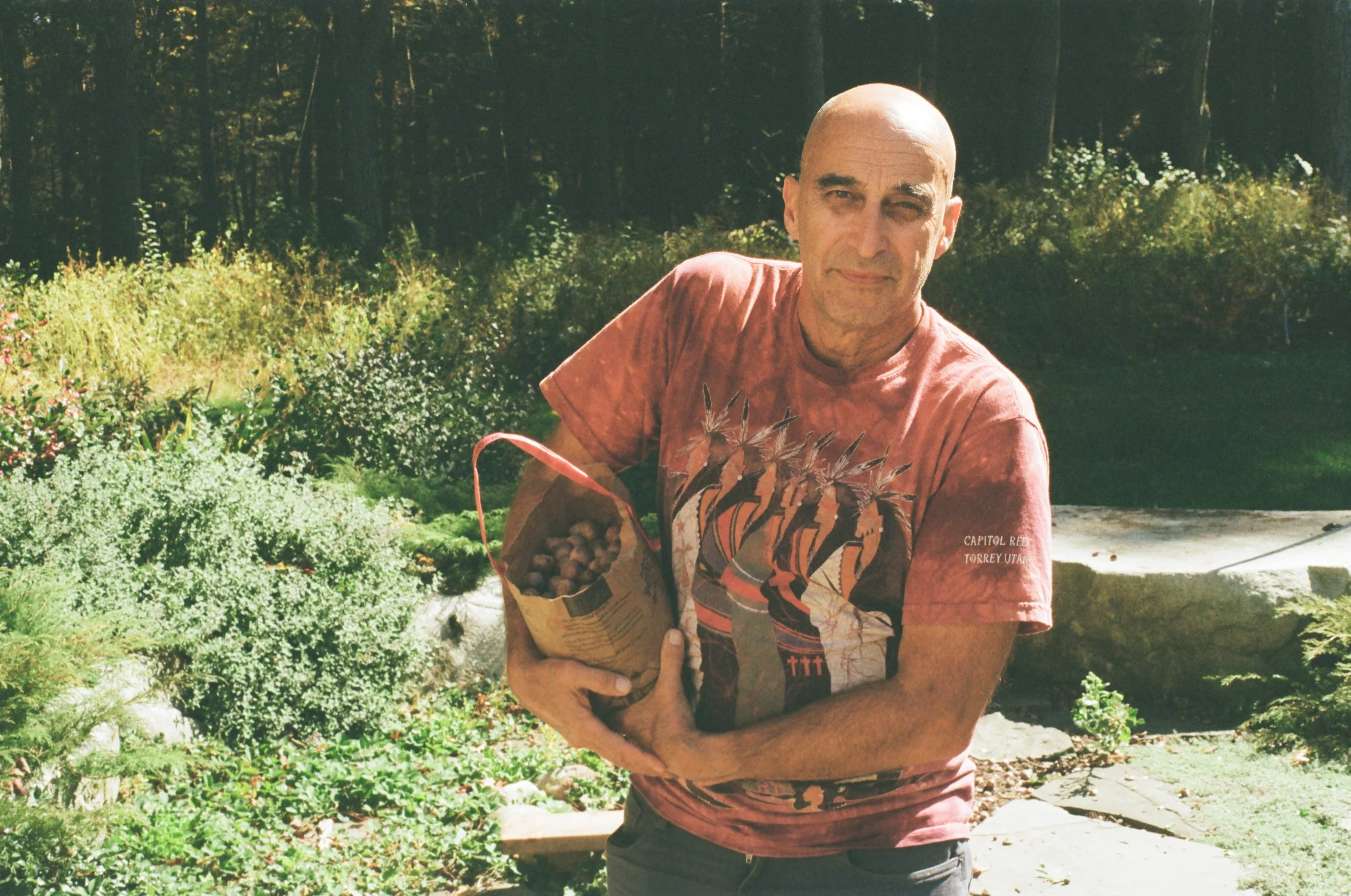 A man with a bald head wearing a red T-shirt holding a paper bag filled with garden produce outdoors.