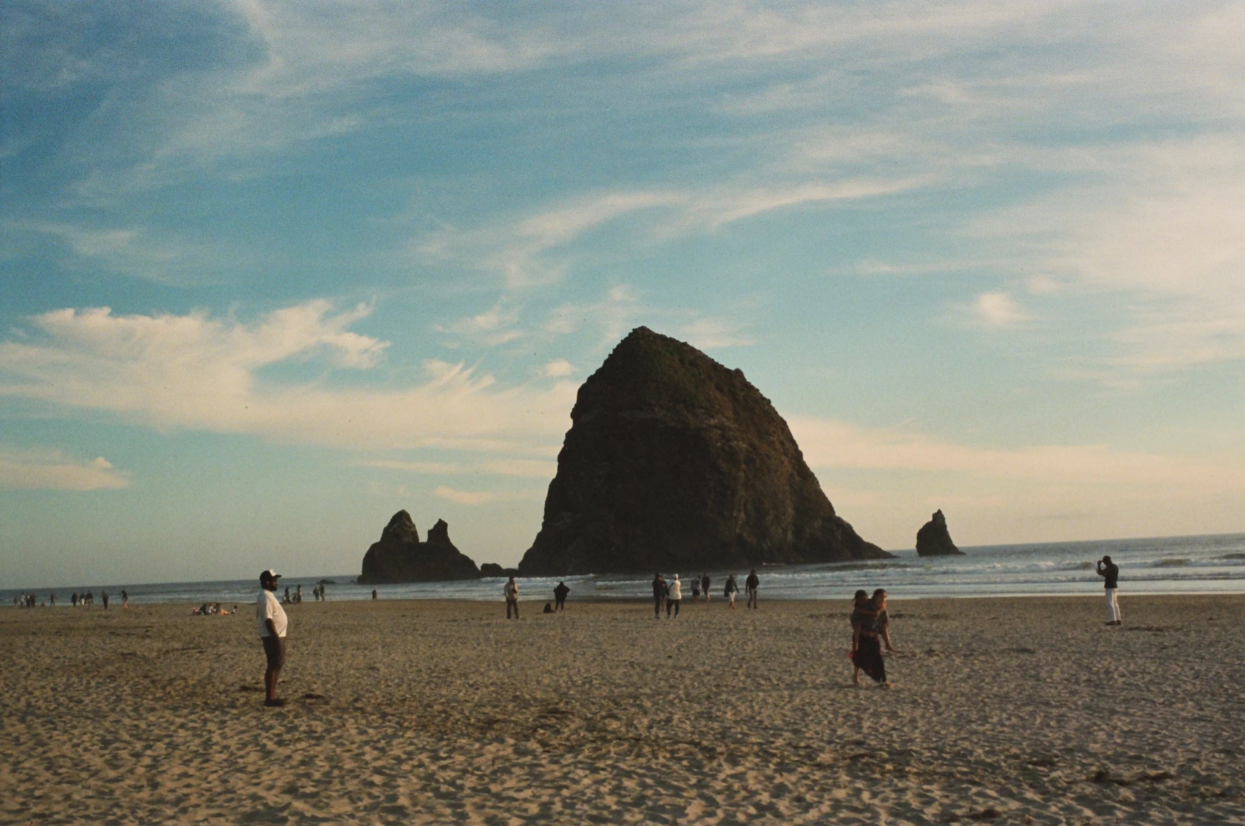 People on a sandy beach near large rock formations in the ocean, with a partly cloudy sky overhead.