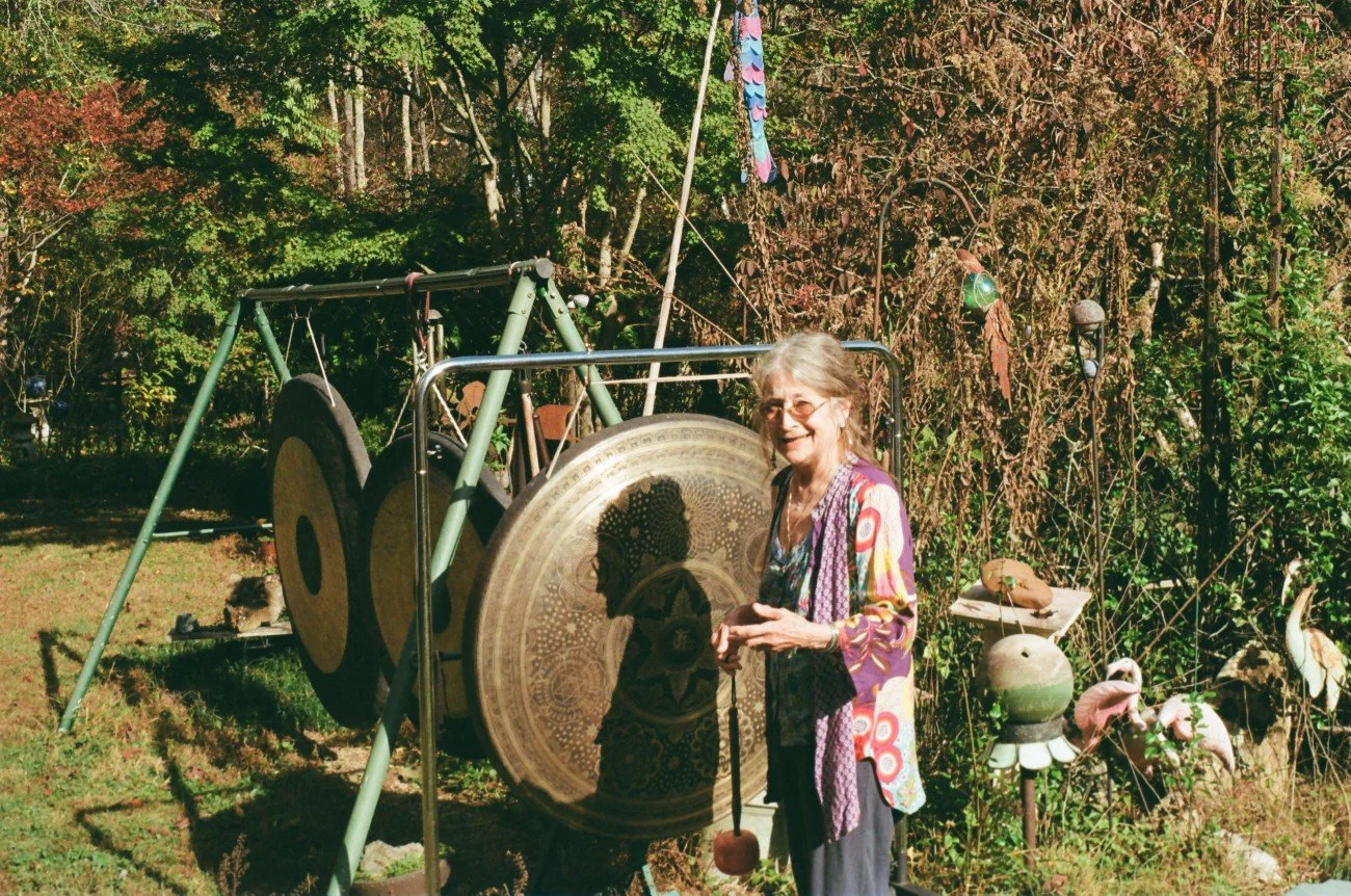 An elderly woman with glasses and long gray hair stands outdoors, smiling, in front of a large Tibetan gong and hung musical instruments. She is wearing a colorful patterned jacket over a dark top and pants. The background features green trees and ga