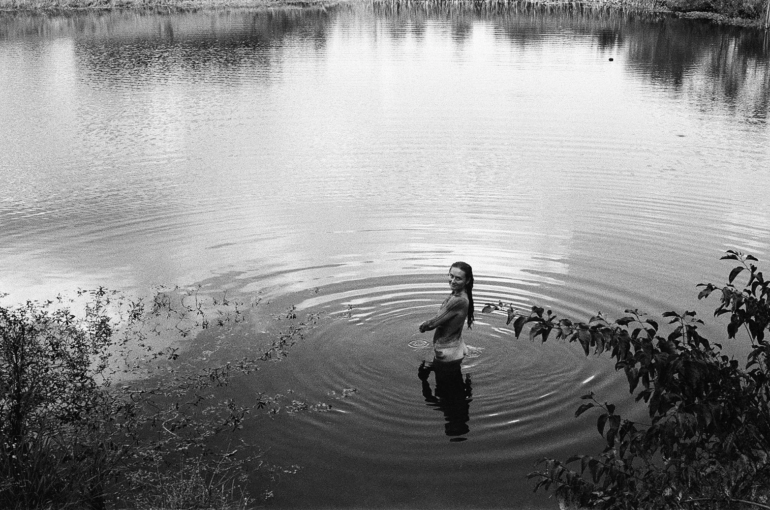 A woman standing waist-deep in a calm body of water, smiling and looking back at the camera, surrounded by trees and bushes, with ripples emanating from her body.