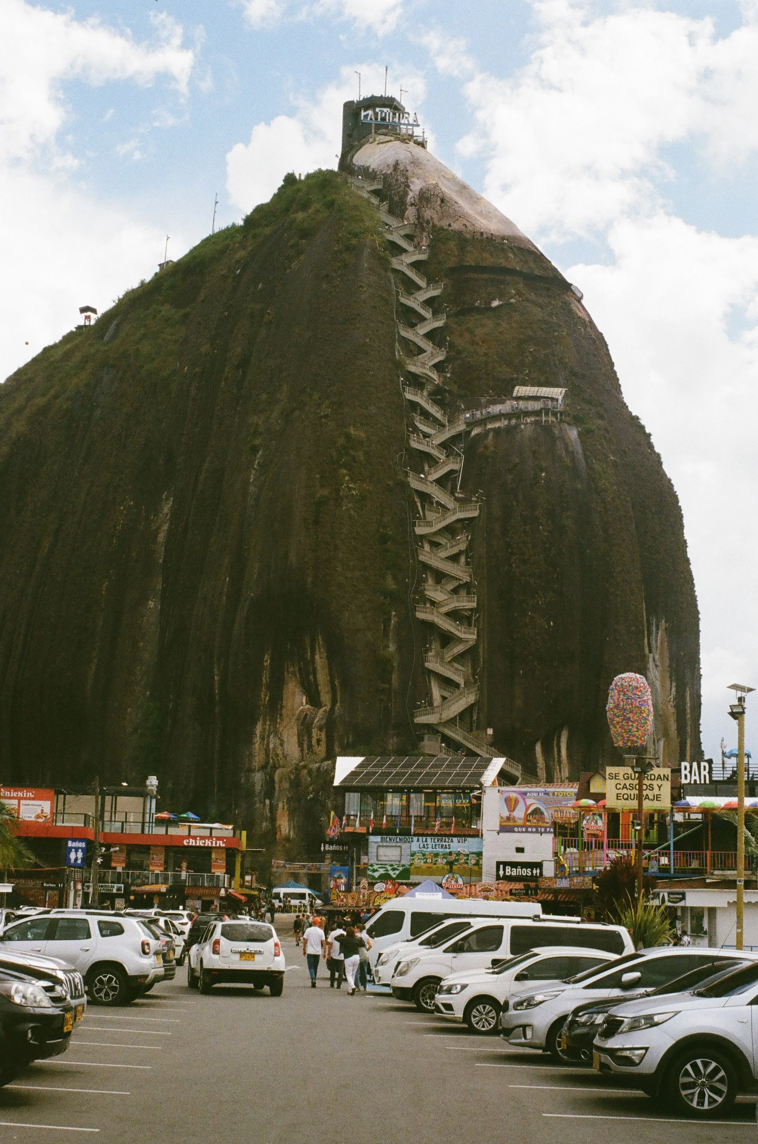 Parking lot with cars and people, in front of a building at the base of a mountain with a zigzag staircase, a sign reading 'La Patera' at the top, and a large egg-shaped sculpture on the right.