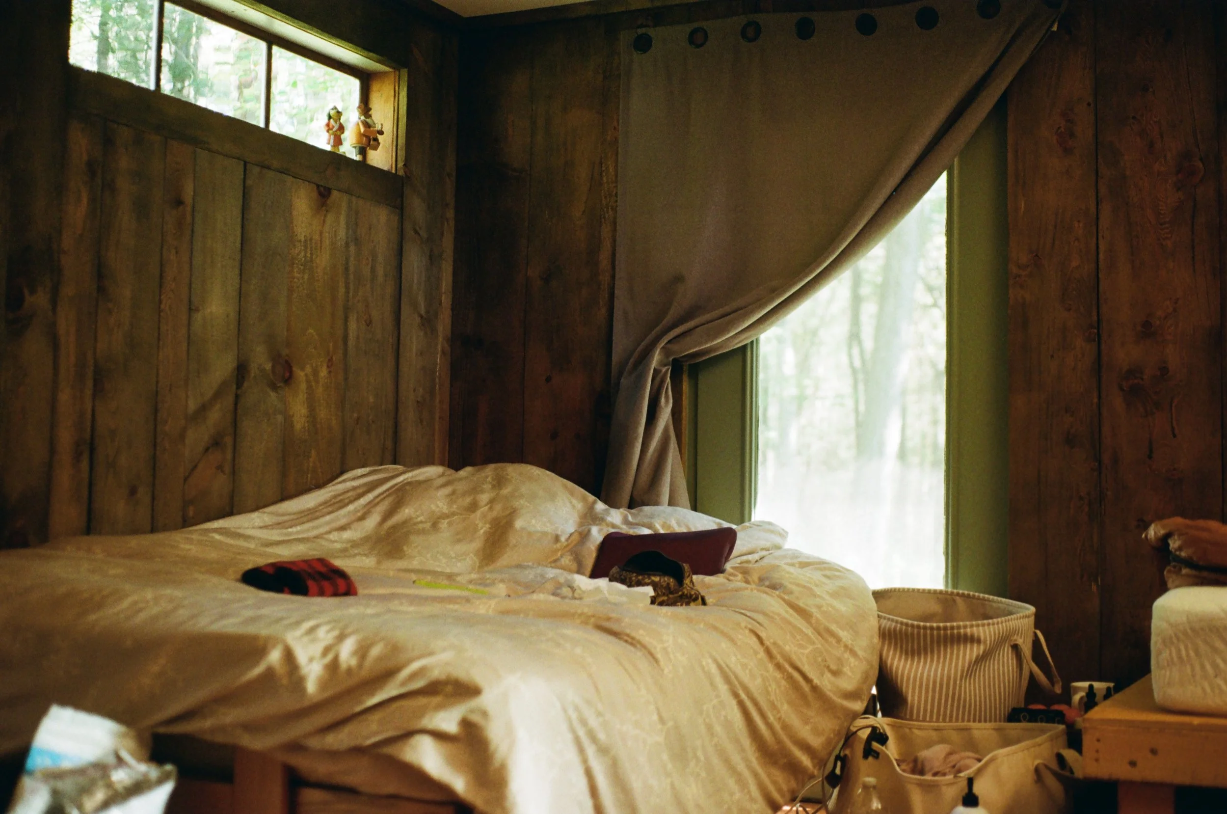 Unmade bed with a cream-colored satin comforter in a wooden-paneled rustic bedroom, with a window behind and a nightstand with various items to the right.