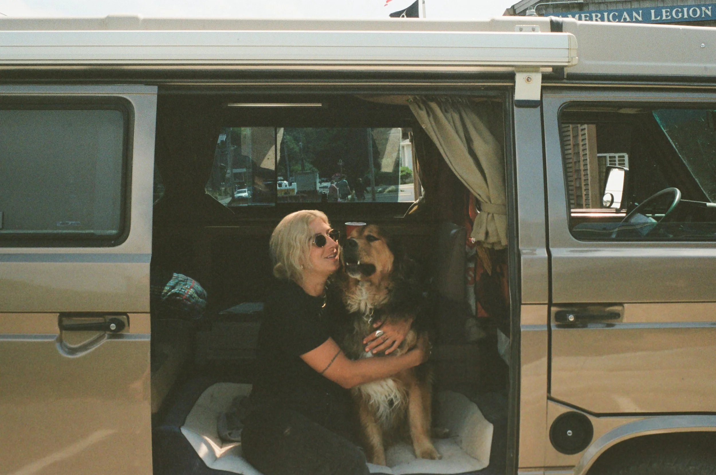 A woman with blonde hair, wearing sunglasses and black clothing, sitting inside a camper van, hugging a large dog, possibly a Bernese Mountain Dog. The camper van has open side doors with curtains, and there are some outdoor elements visible through 