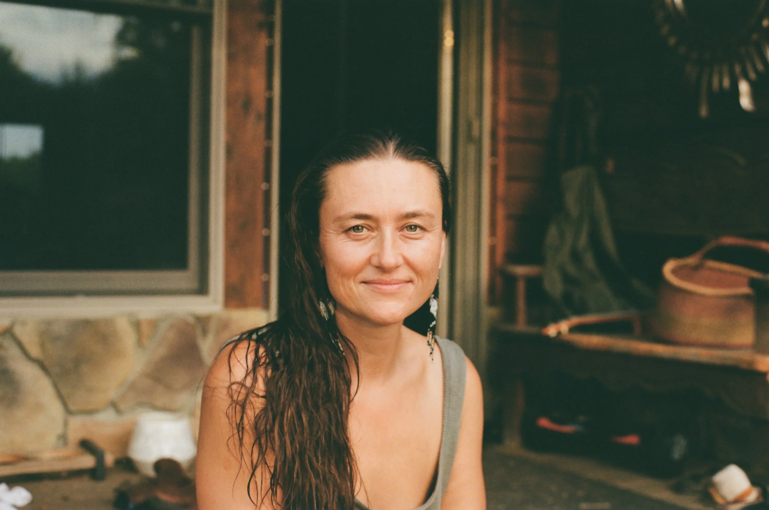 A woman with long dark hair and earrings, wearing a tank top, standing outside near a house with a window, stone wall, and wooden deck with outdoor furniture and a bag in the background.
