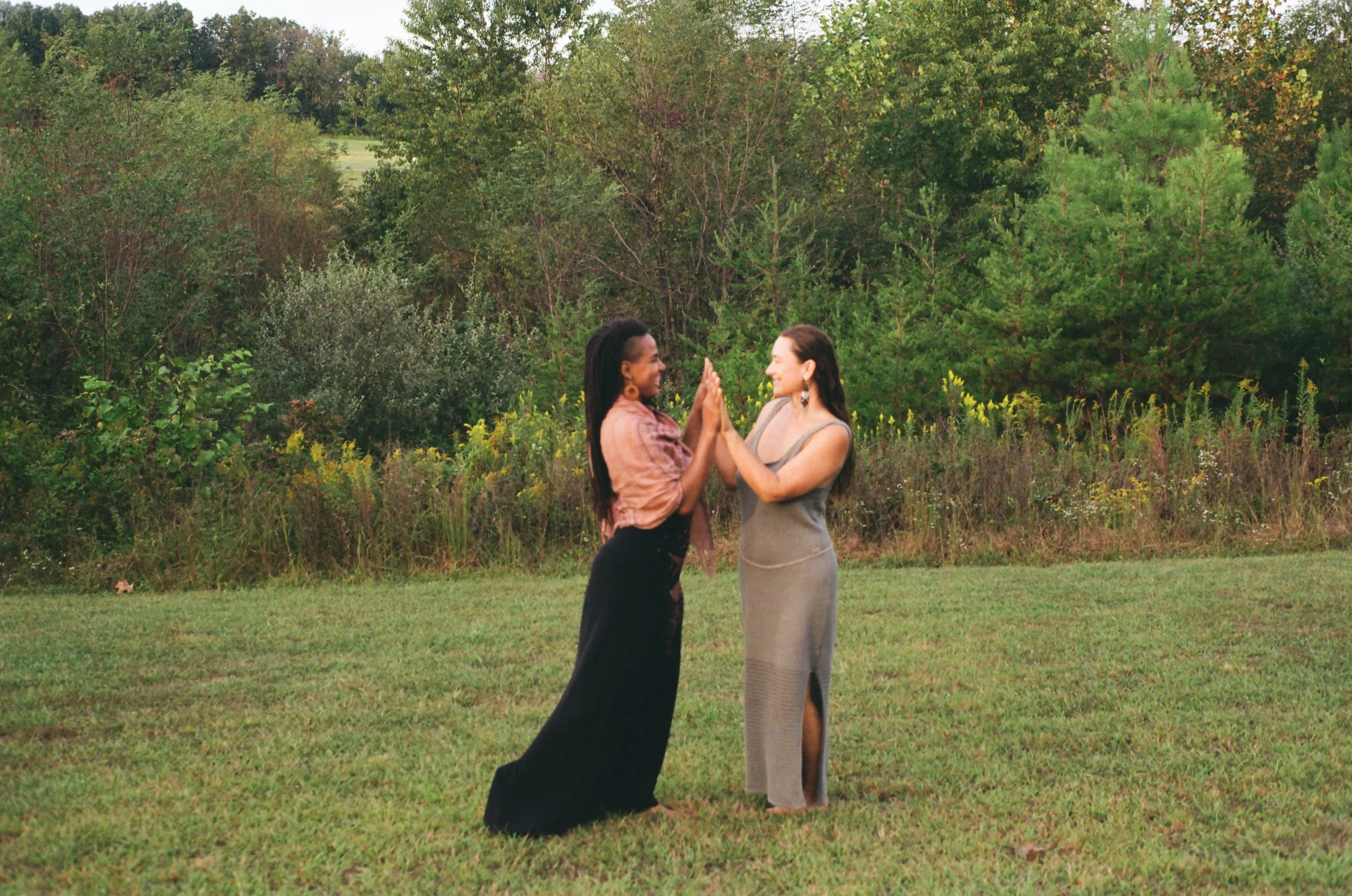 Two women standing in a grassy field with trees in the background, touching hands and smiling at each other.