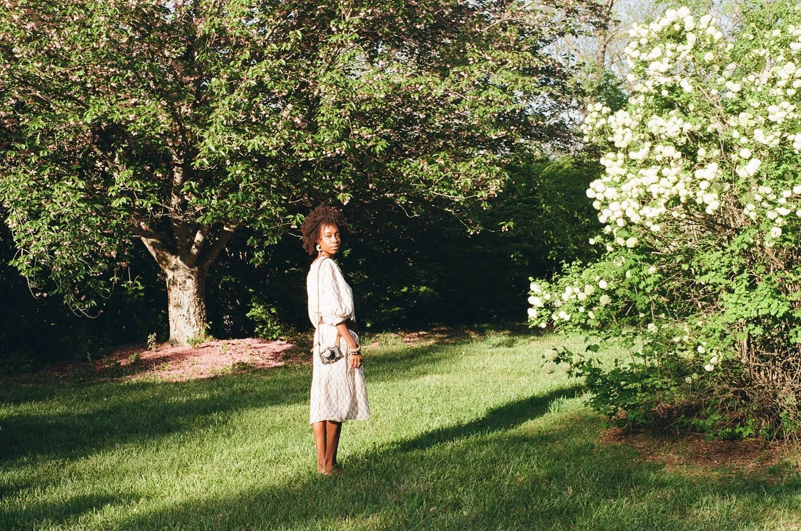 A woman in a long, patterned dress stands barefoot on green grass in a garden, holding a small camera, with trees and white flowering bushes in the background.