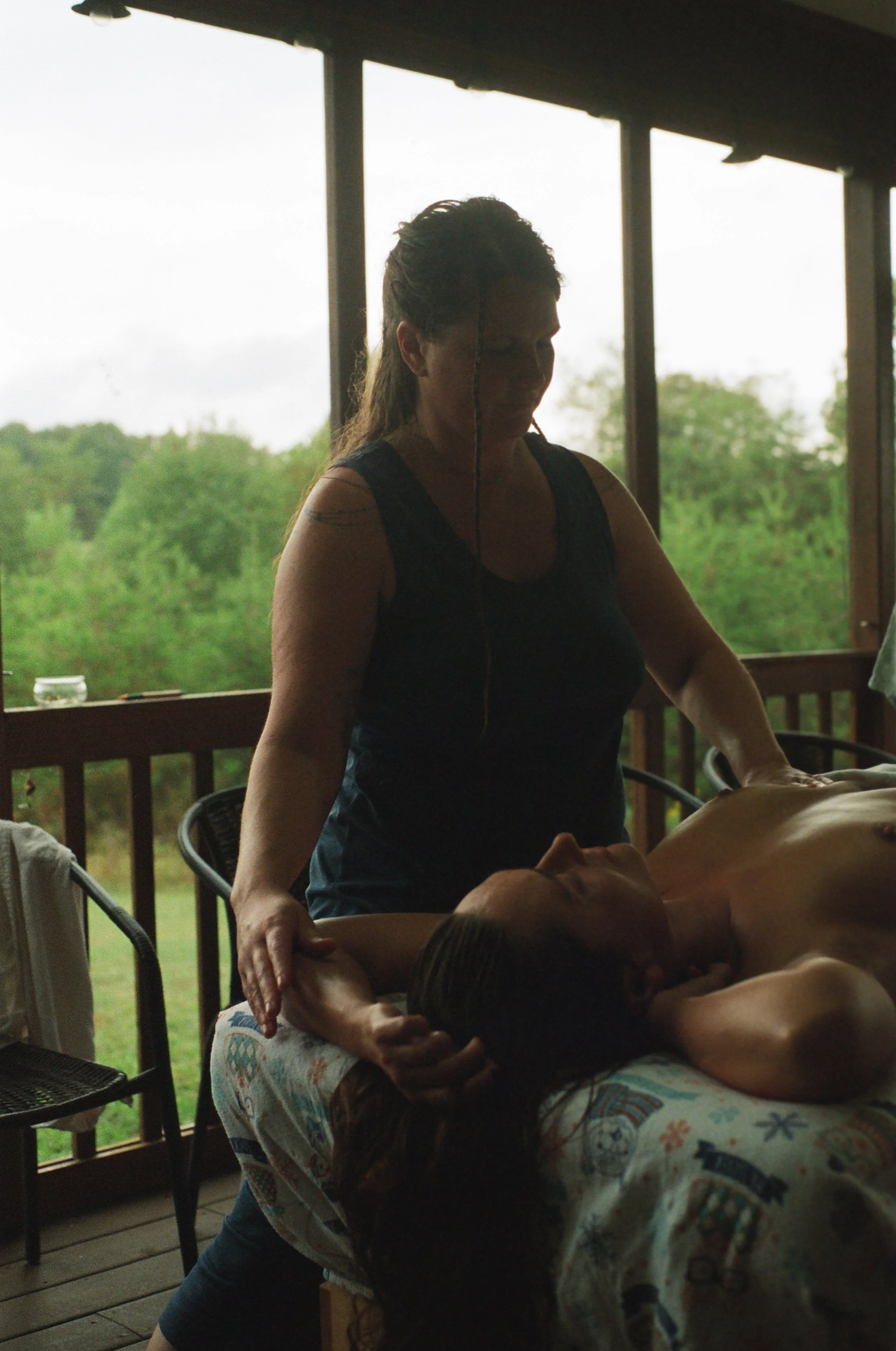 A person giving a massage to another person lying on a table, with a view of green trees outside the window.