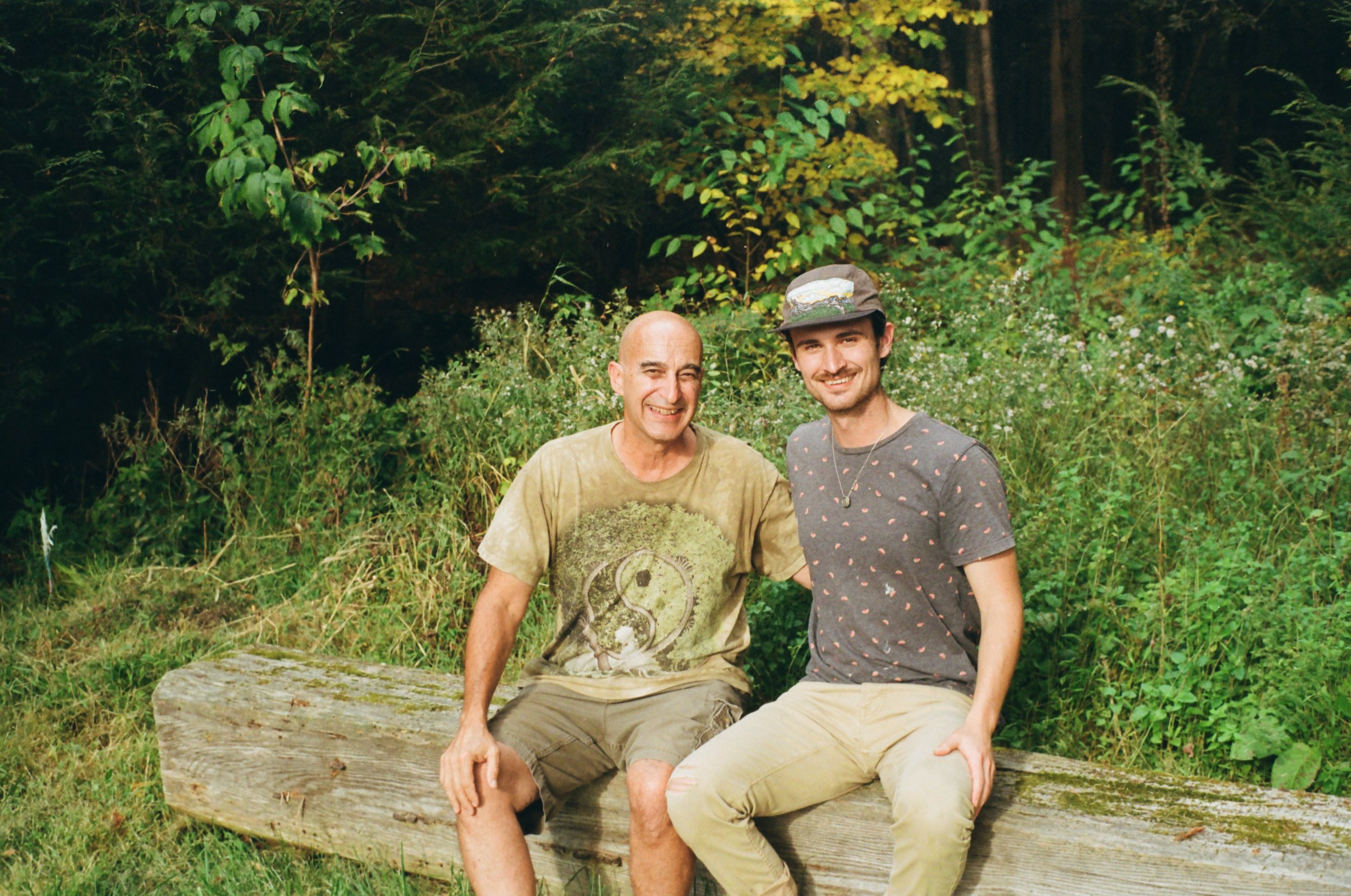 Two men sitting on a wooden log in a green, wooded outdoor setting, smiling at the camera.