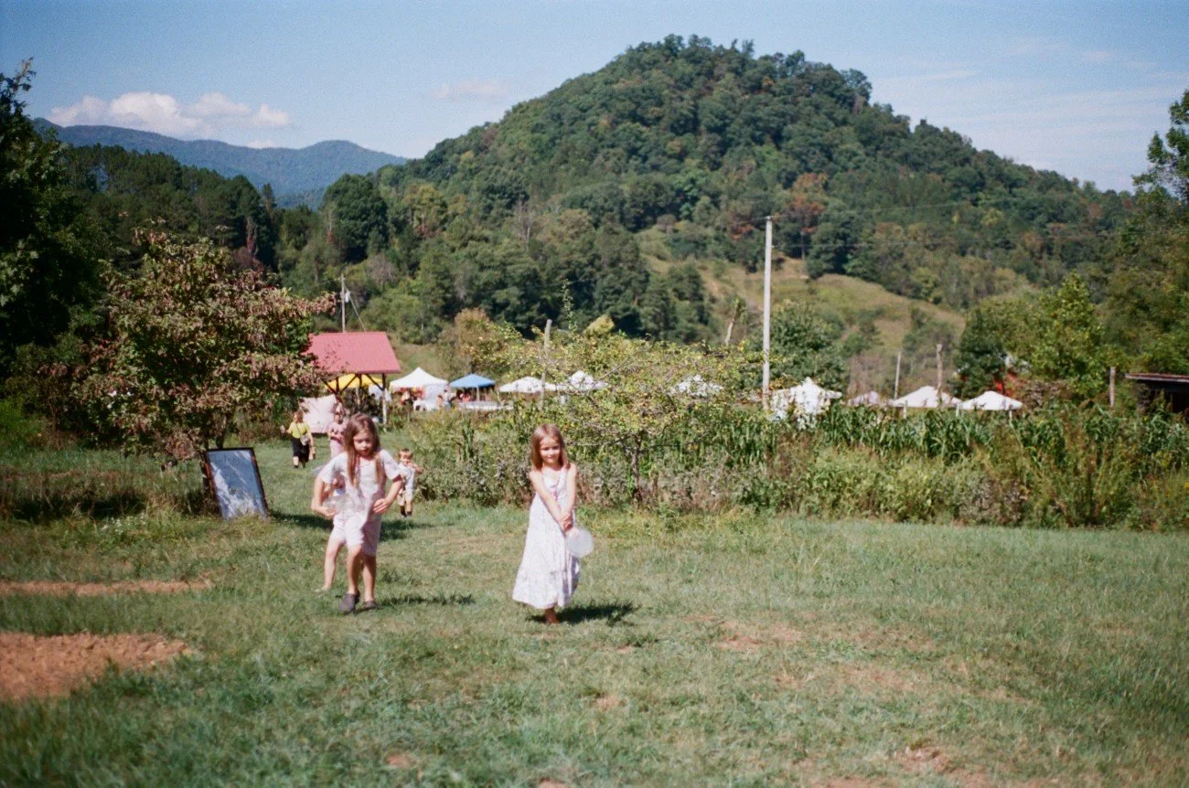 Children playing on a grassy field with trees, hills, tents, and a small building in the background, under a partly cloudy sky.