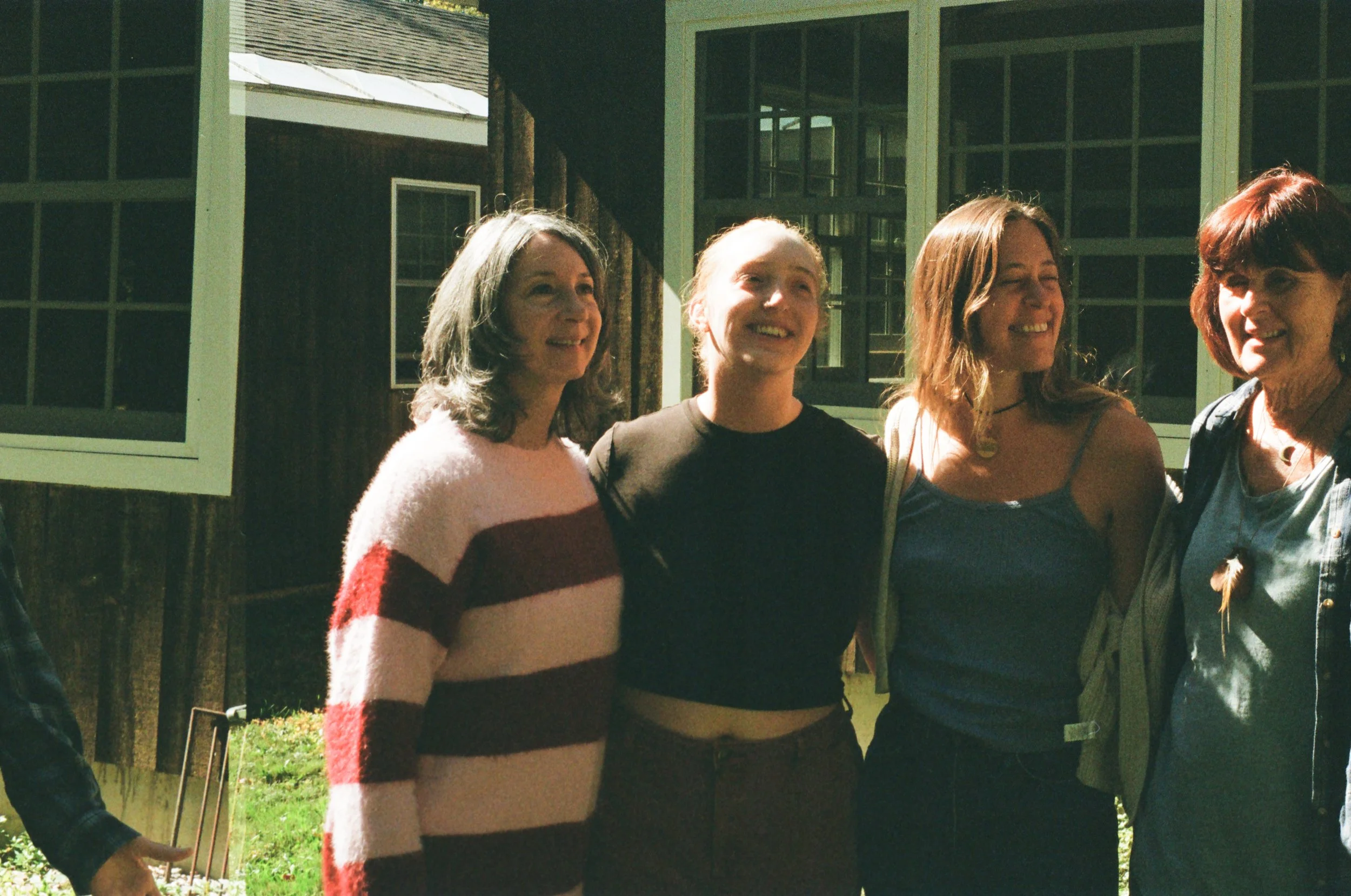 Four women standing outdoors in front of a wooden building with green window frames, smiling and enjoying a sunny day.