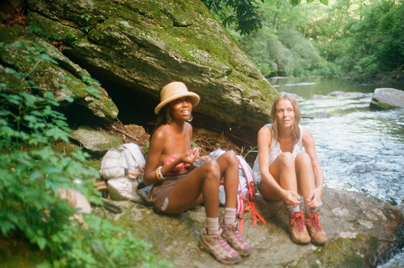 Two women sitting on a rock by a river in a lush green forest, with outdoor hiking gear and backpacks.