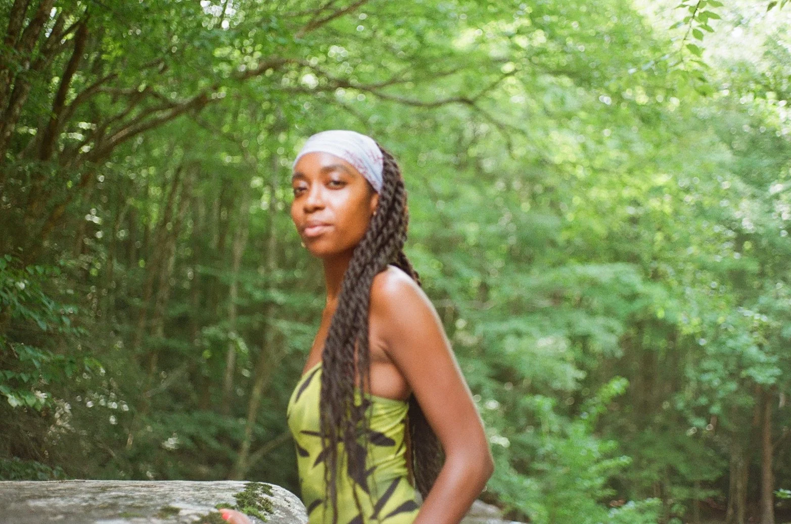 A young woman with long braids and a light-colored headscarf standing outdoors in a lush green forest, wearing a sleeveless green top, facing slightly to the left.