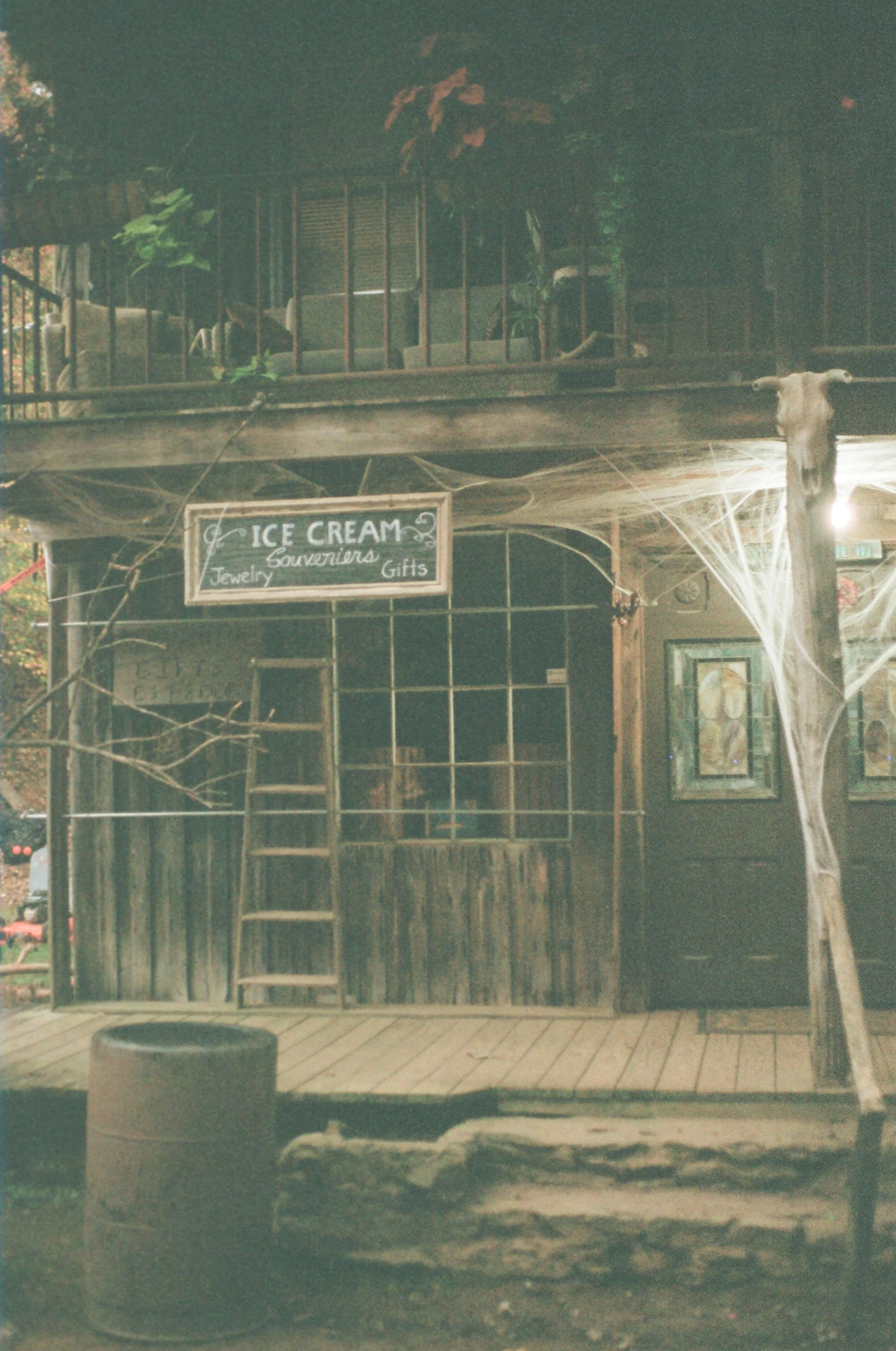 Old wooden storefront with a sign reading 'Ice Cream, Souvenirs, Jewelry, Gifts,' decorated with cobwebs, in a rustic setting with a staircase and artwork on the wall.