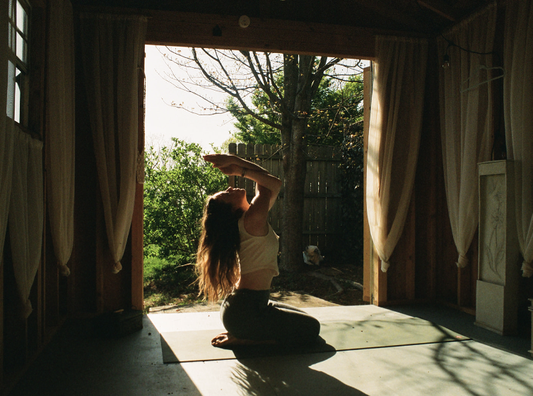 Woman practicing yoga on a mat in a sunlit room with open doors leading to a backyard with trees.