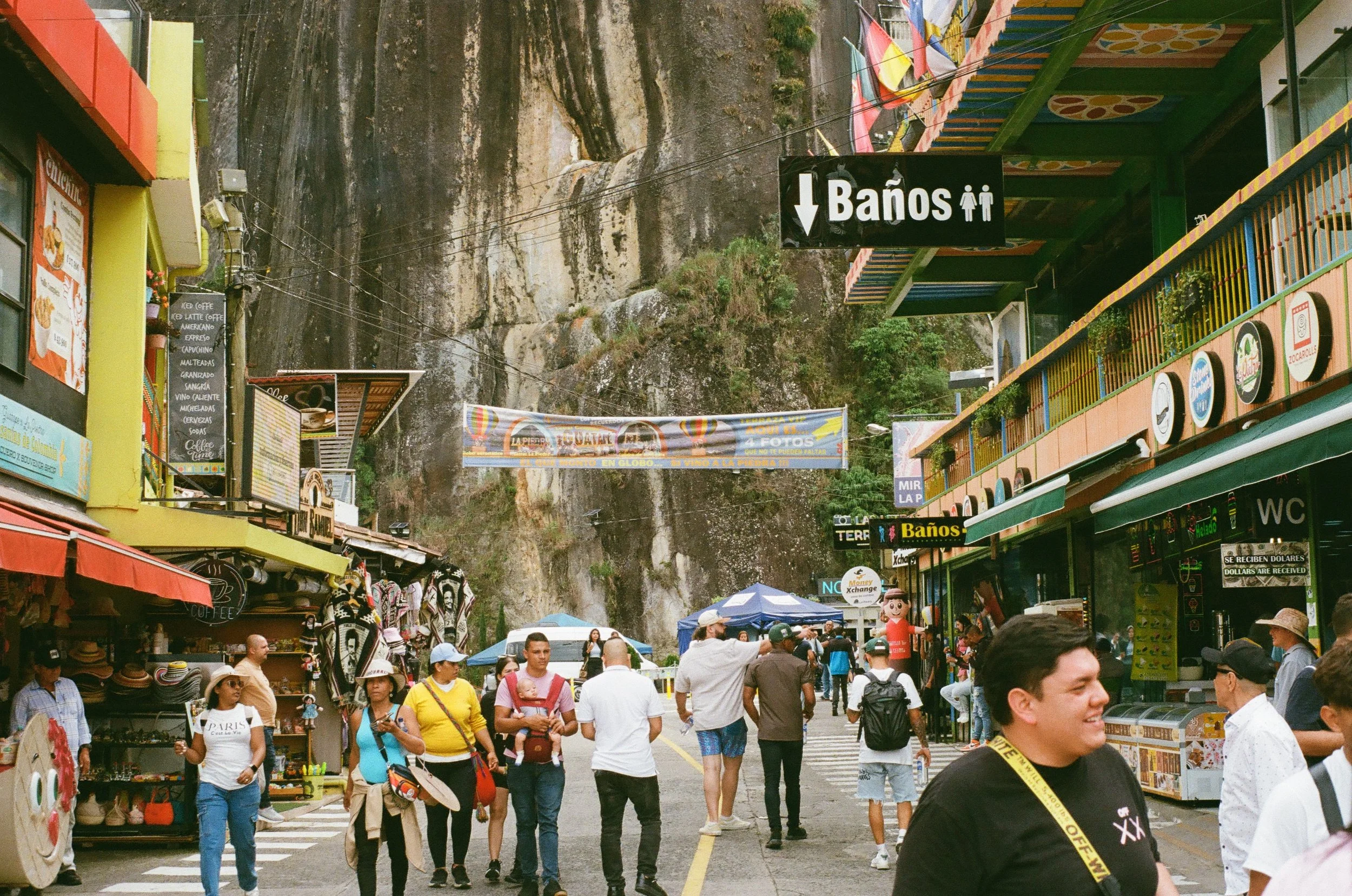 A busy street scene in a tourist area with many people walking and shopping. There are colorful shops, signs in Spanish, and a large rock face in the background. A sign indicates the direction to restrooms.