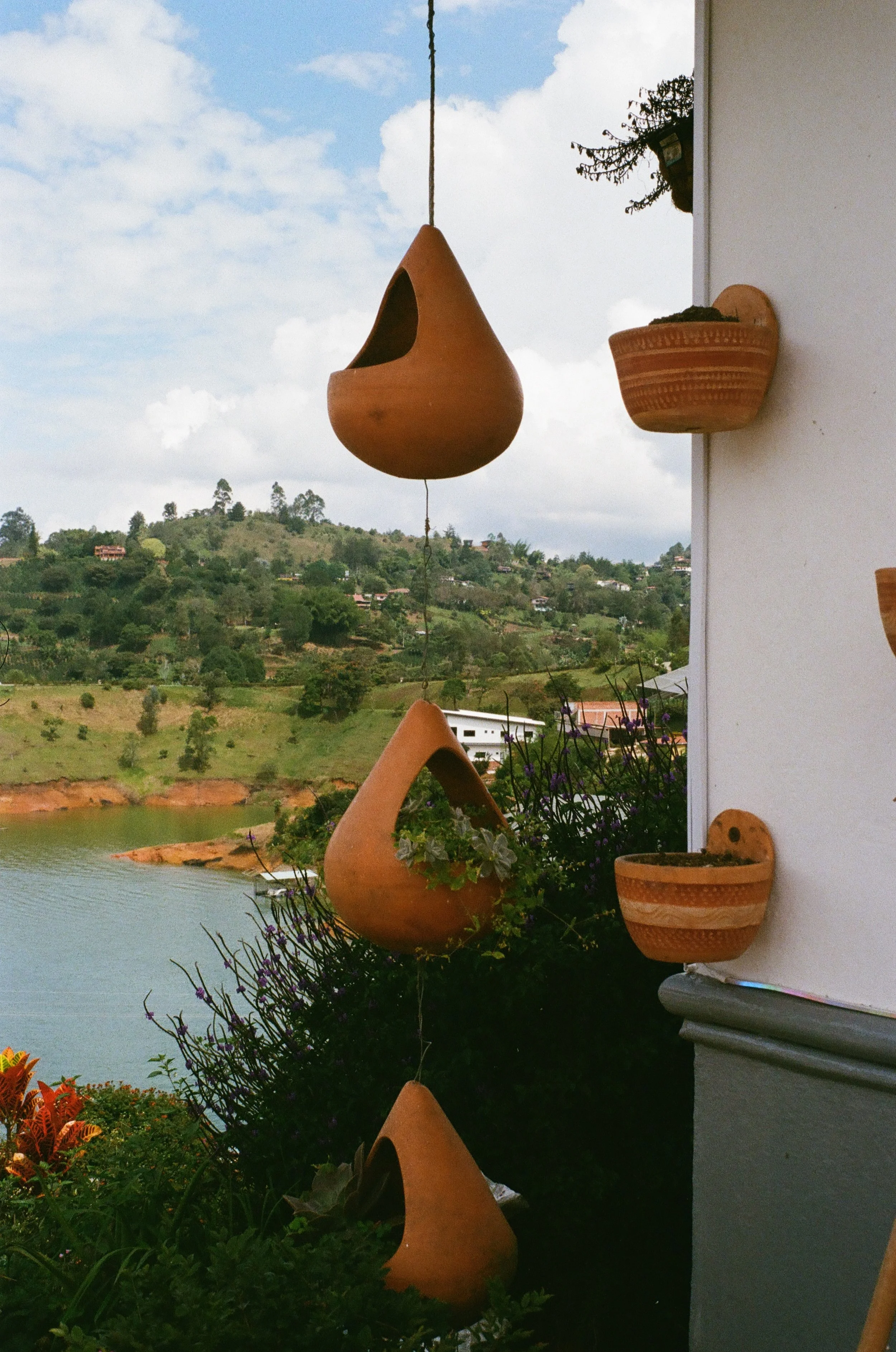 Terracotta hanging planters and wall-mounted pots on a balcony with a scenic landscape of a lake, hills, and houses in the background.