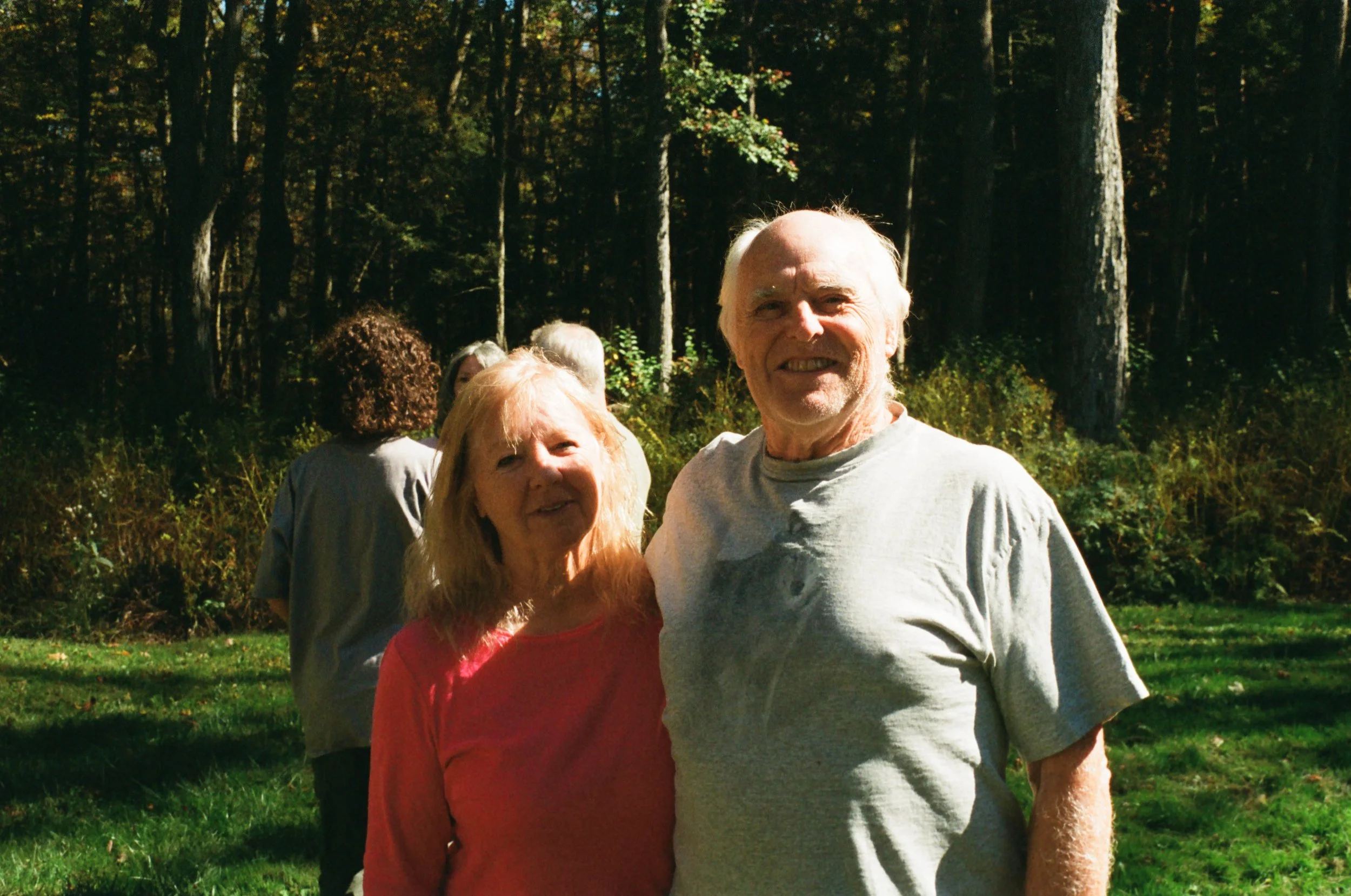 Senior couple standing outdoors in a grassy area with trees in the background, smiling at the camera.