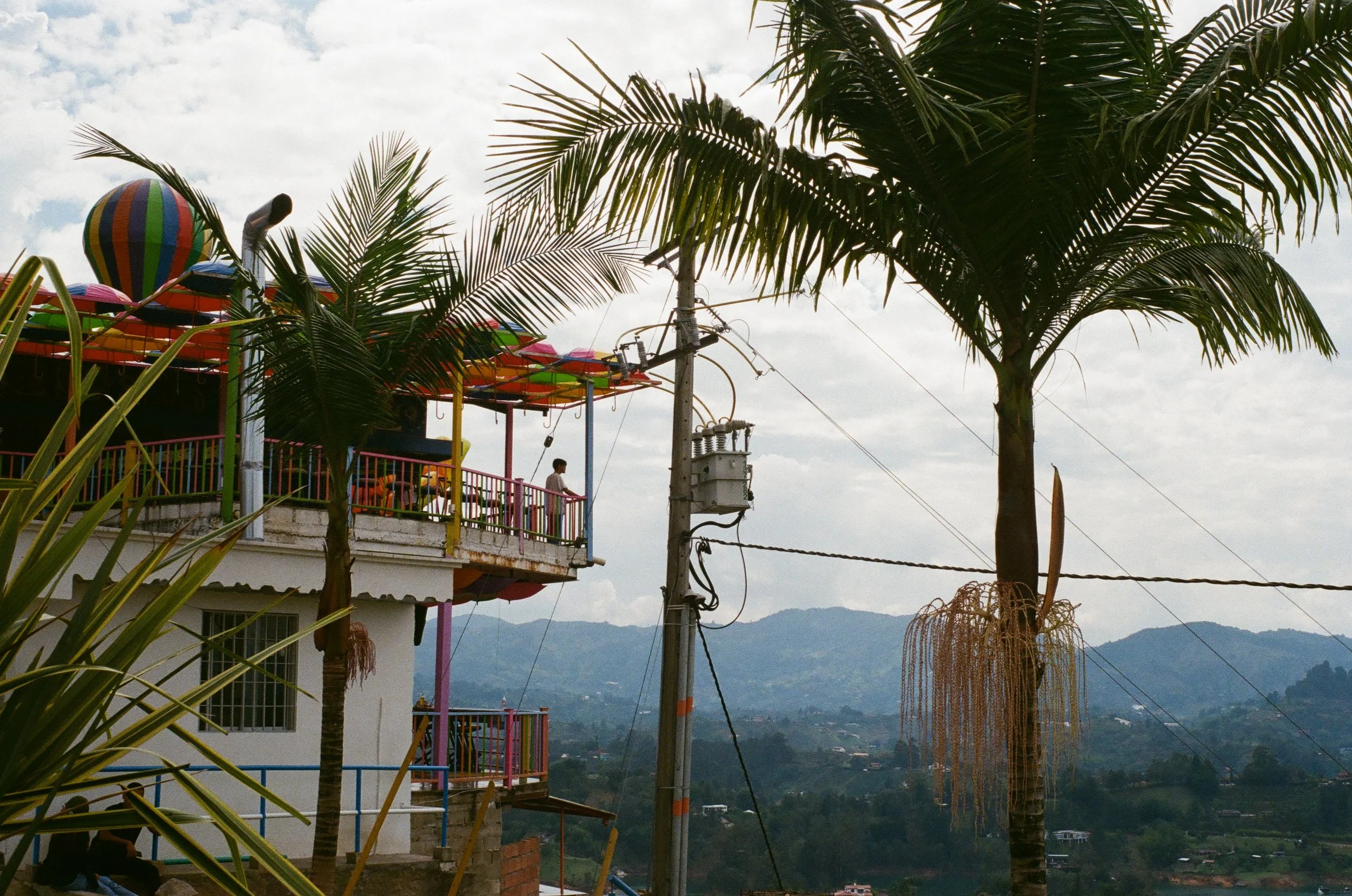 A colorful amusement park ride with a rainbow-striped hot air balloon and a slide, surrounded by palm trees, an electric pole, and a mountain landscape in the background.