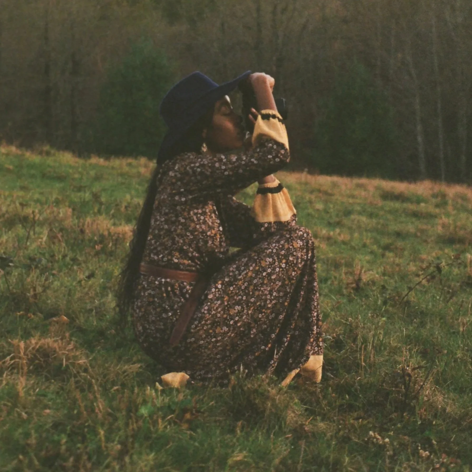 Woman in floral dress and wide-brimmed hat kneeling on grass, looking through a camera in a field during sunset or dusk.