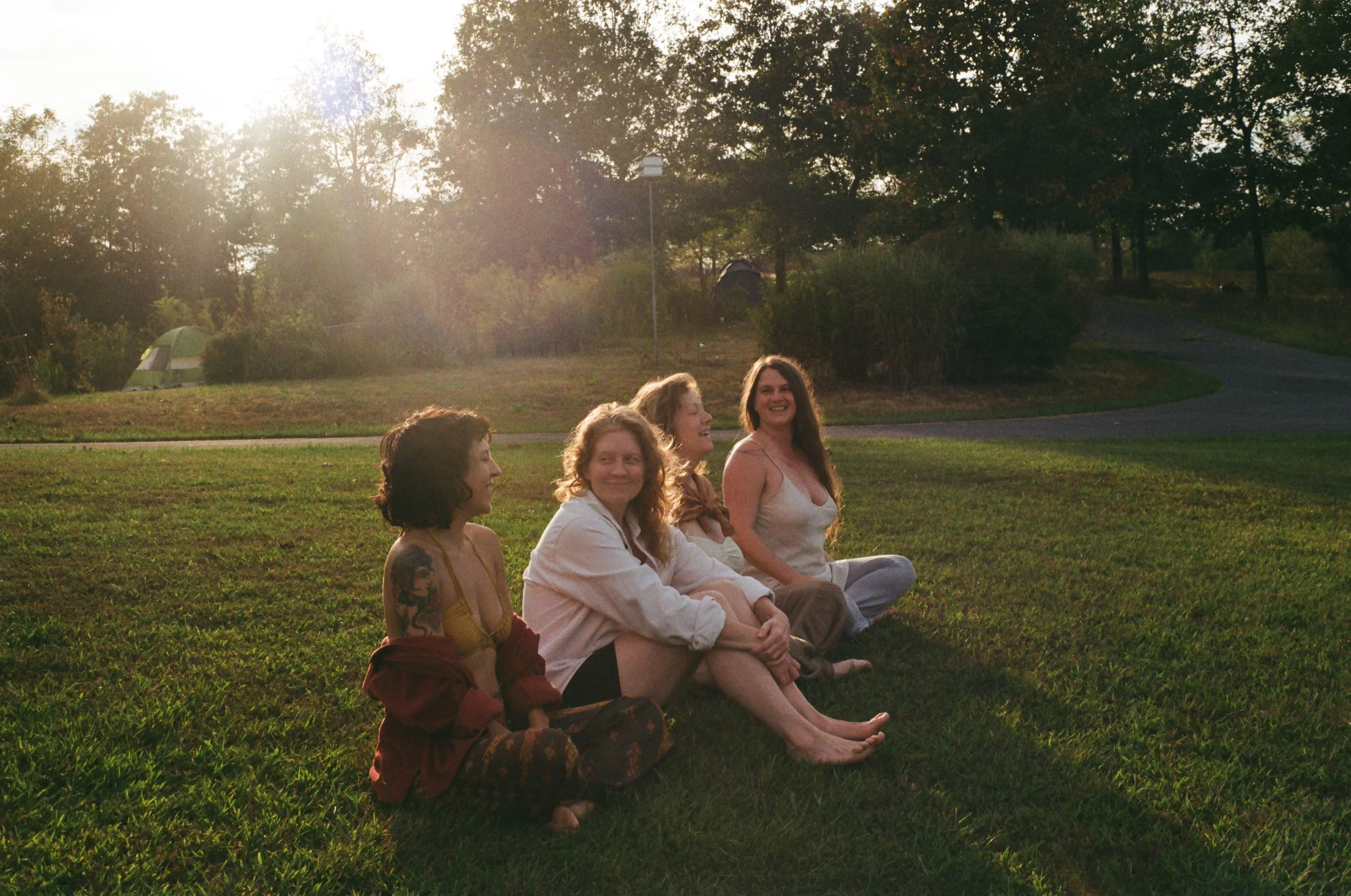 Four women sitting on grass in a park with trees, a tent, and a lamppost in the background during sunset, smiling and enjoying each other's company.