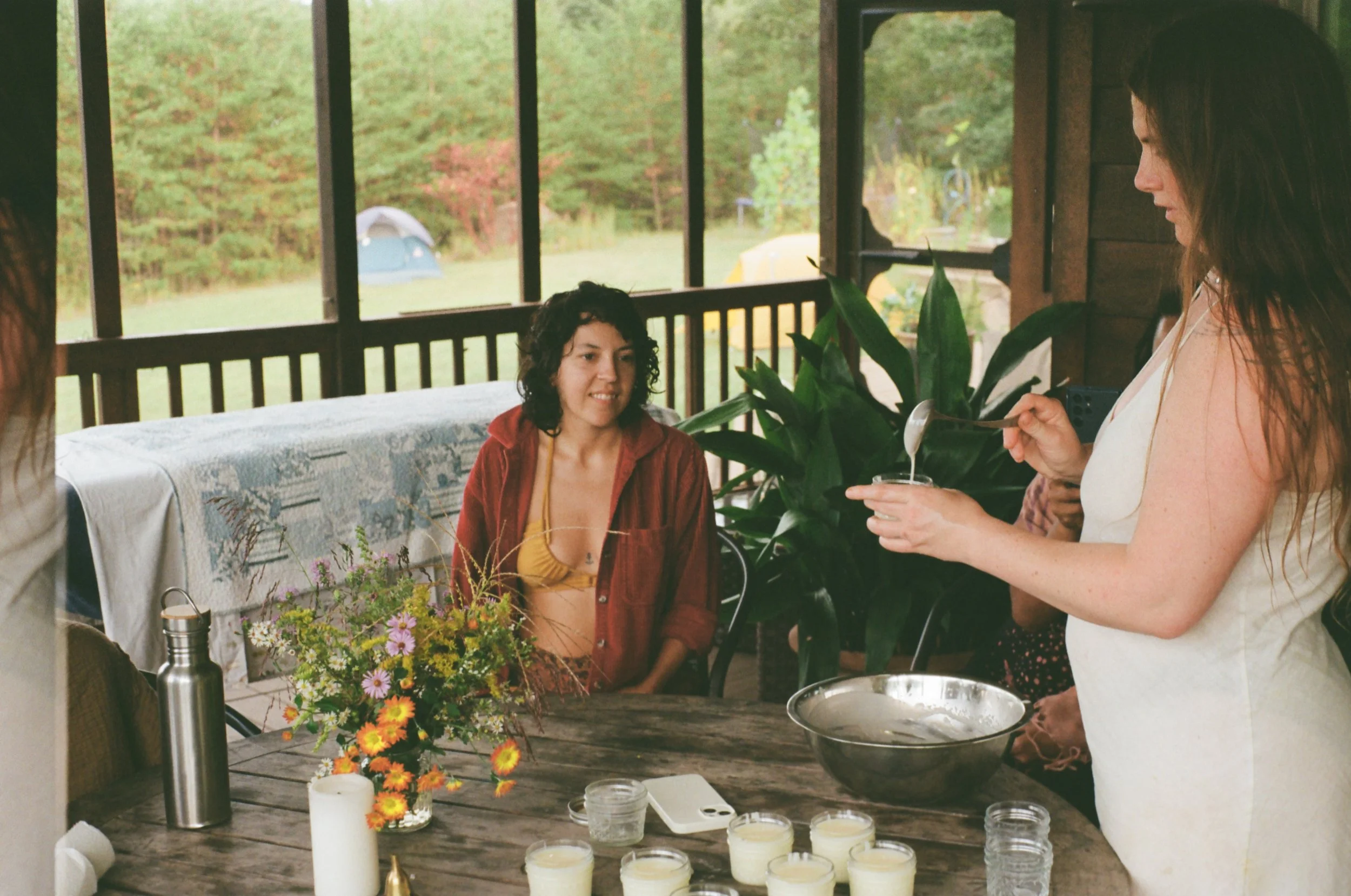 People gathered around a table on a porch, with one woman pouring a creamy mixture into a bowl while others observe, decorated with candles and a flower arrangement, overlooking a grassy yard and trees outside.
