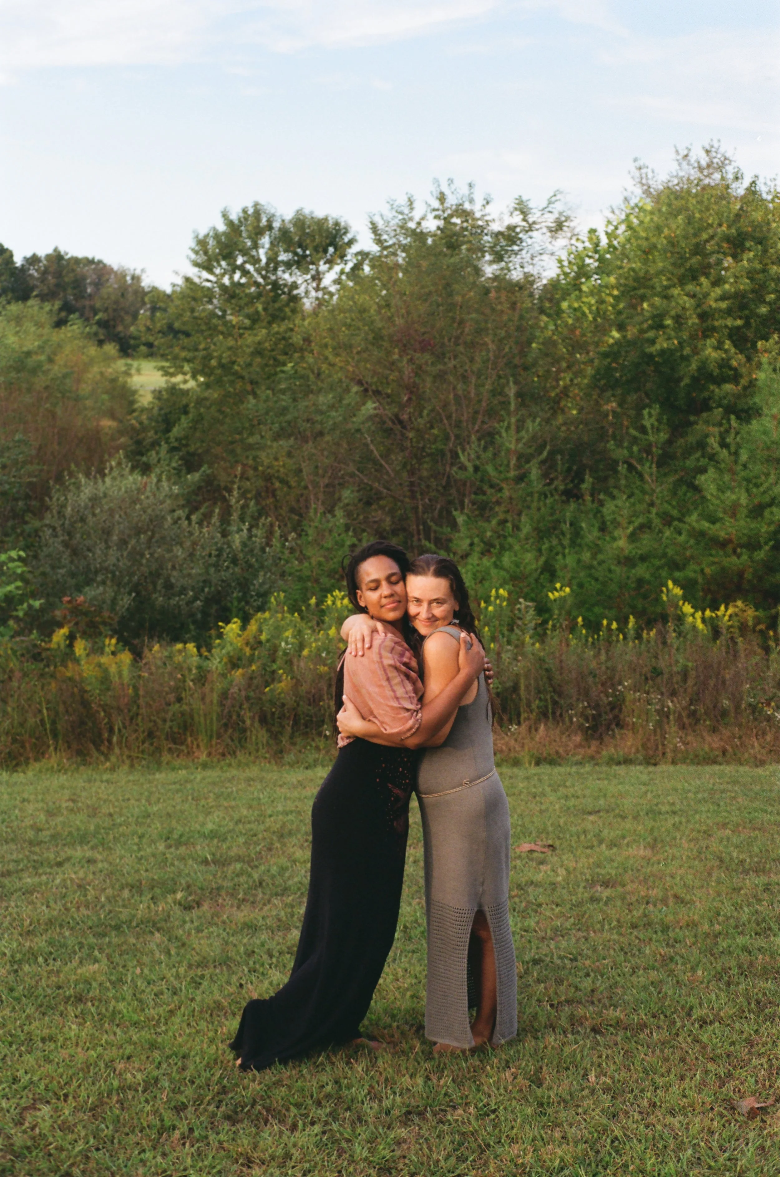 Two women hugging outdoors on a grassy field with trees and a blue sky in the background.