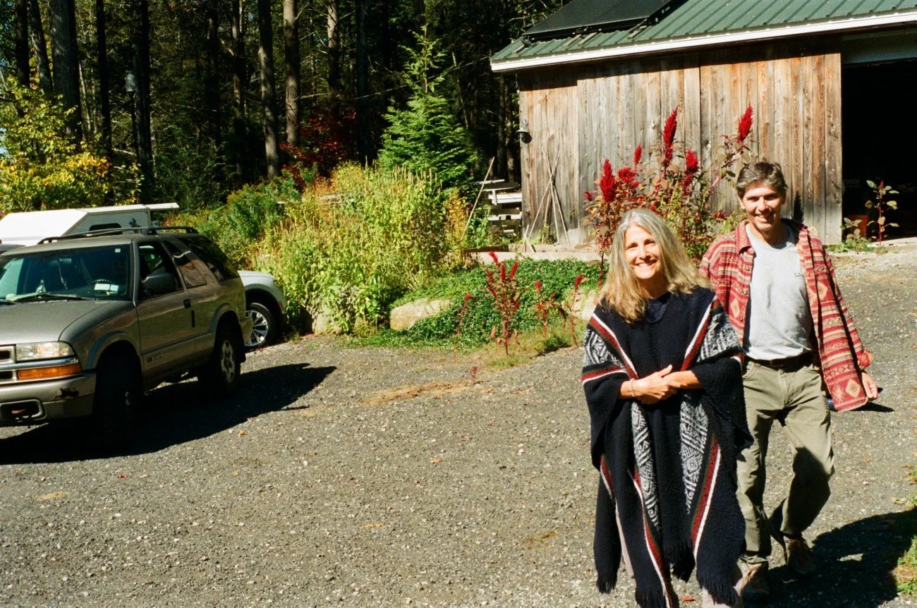 Two people walking on a gravel driveway in front of a wooden building with plants and a forest in the background.