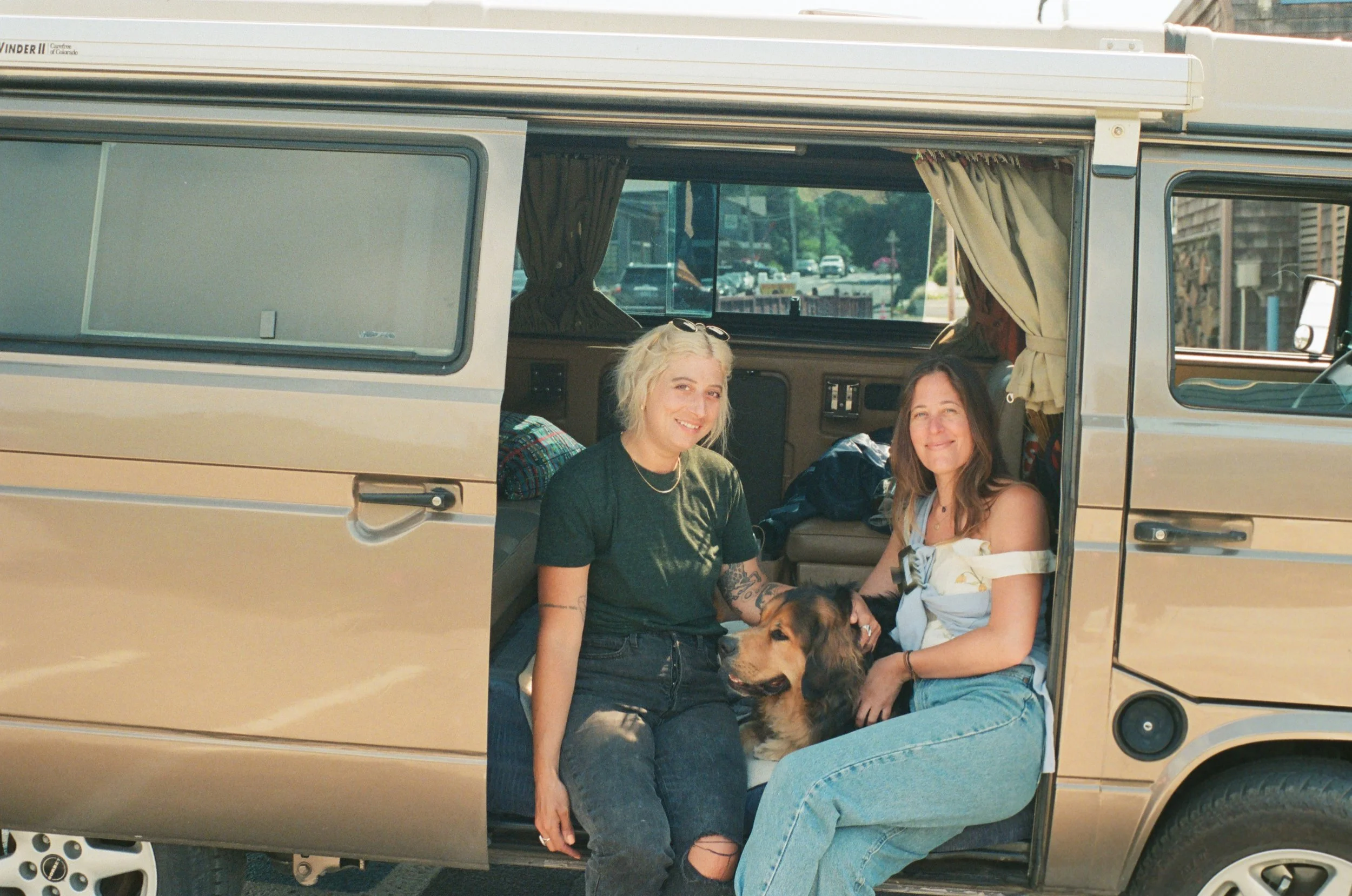 Two women sitting inside a vintage van with a dog between them, in a parking lot.