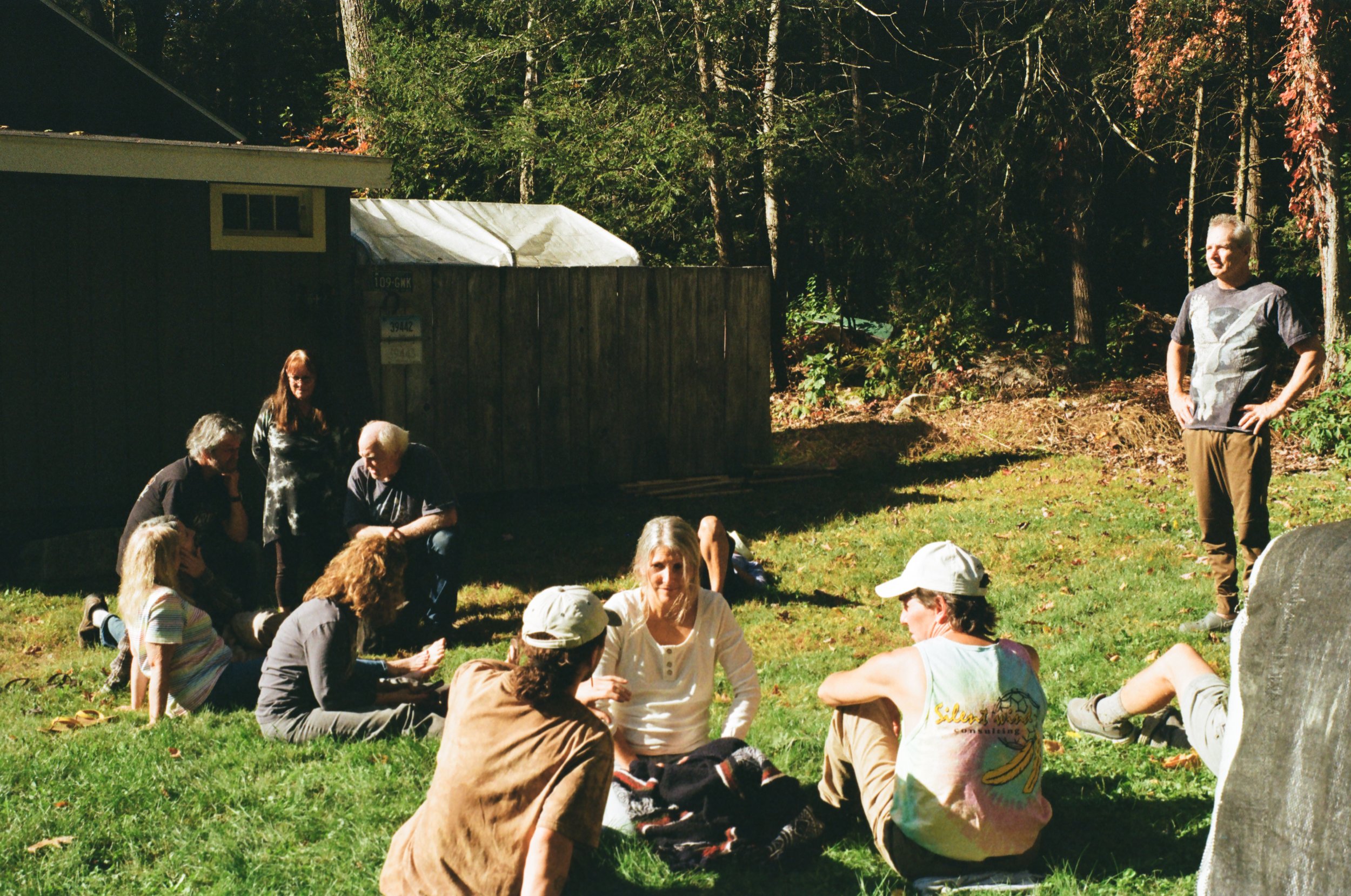 A group of people sitting and standing on a grassy yard, with a wooden fence and trees in the background, enjoying a sunny day.