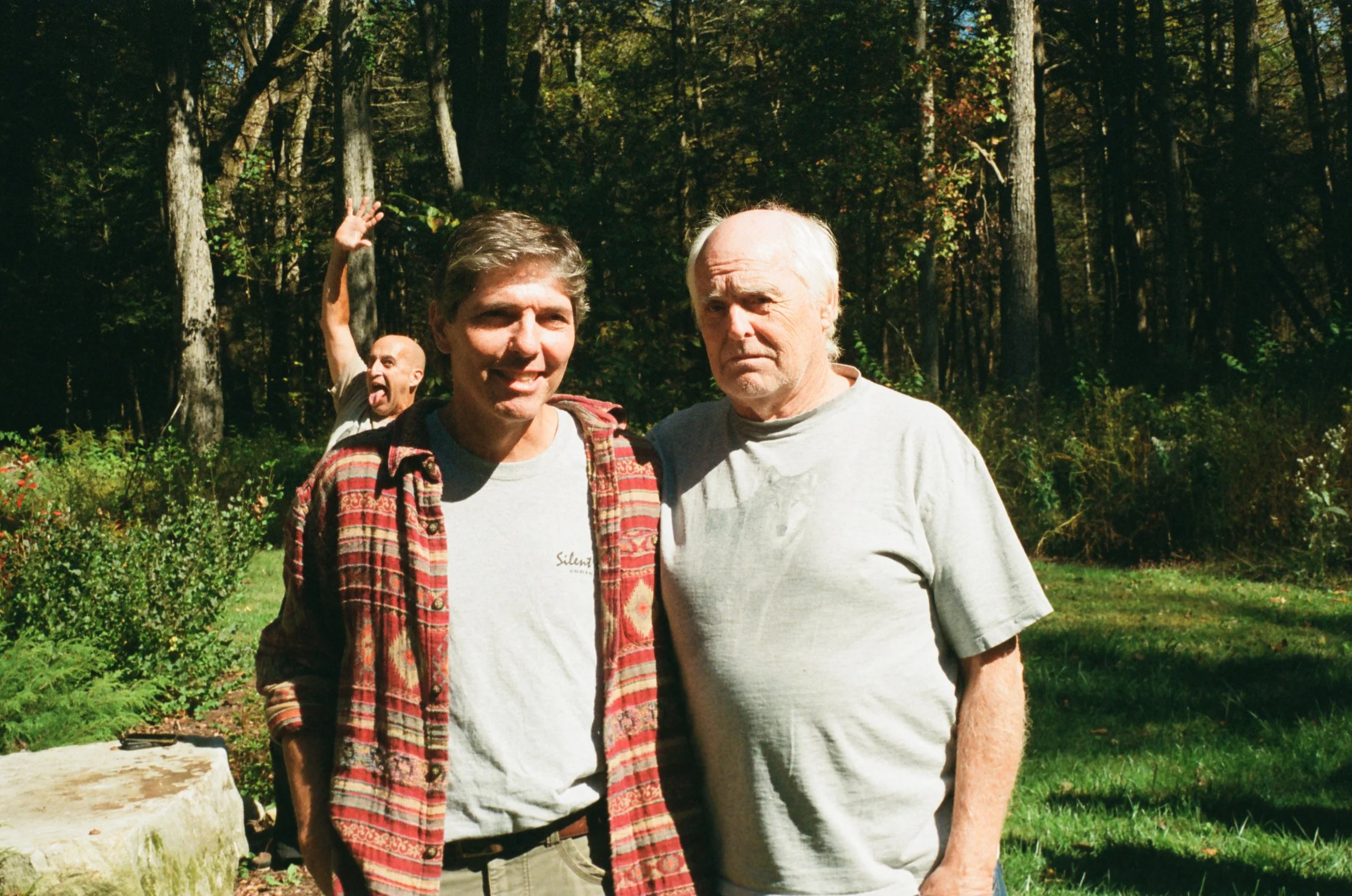 Two men standing outdoors in a forested area, one older with white hair and one younger with brown hair, both wearing casual t-shirts. A third man in the background is making a funny face and raising his hand.
