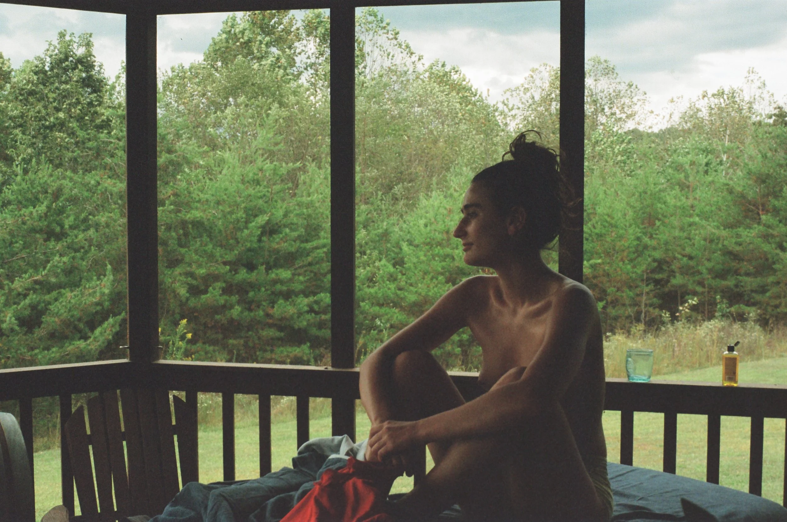 A woman with dark hair tied up, sitting on a bed in a screened porch, looking to the right with a smile. Outside the porch, there are green trees and a cloudy sky. Items like a blue glass, a yellow bottle, and some clothing are on the bed.