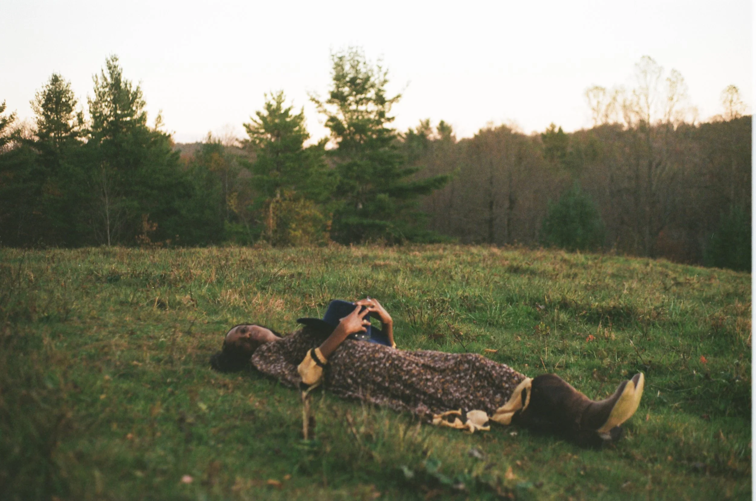 Person lying on the grass in a rural area during sunset, holding a hat and wearing a patterned coat and boots.