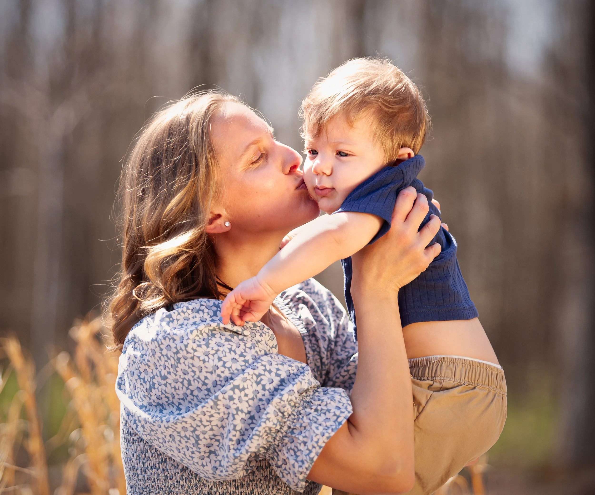 A woman with wavy, light brown hair holding and kissing a young boy with light brown hair and blue eyes, outdoors in a natural setting with blurred trees and plants in the background.