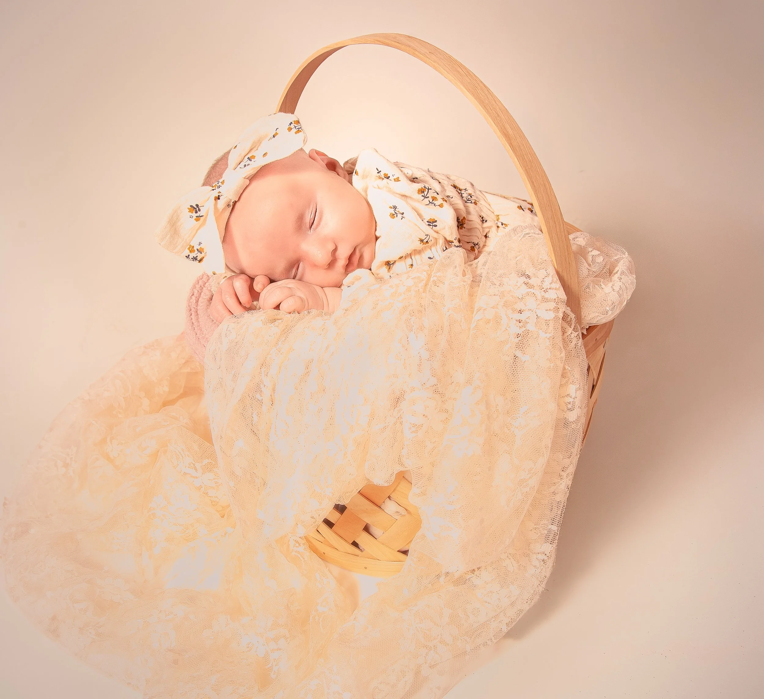 A sleeping baby with a headband and floral outfit, laying on a lace-covered surface with a wooden decorative arch.