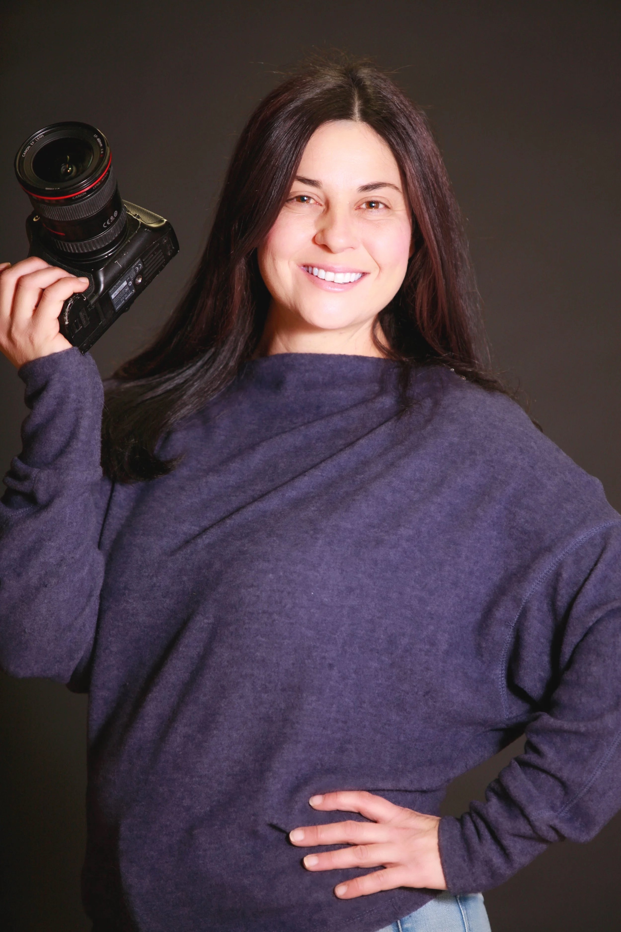 A woman with long dark hair smiling and holding a camera on her shoulder, wearing a dark purple sweatshirt, against a dark background.