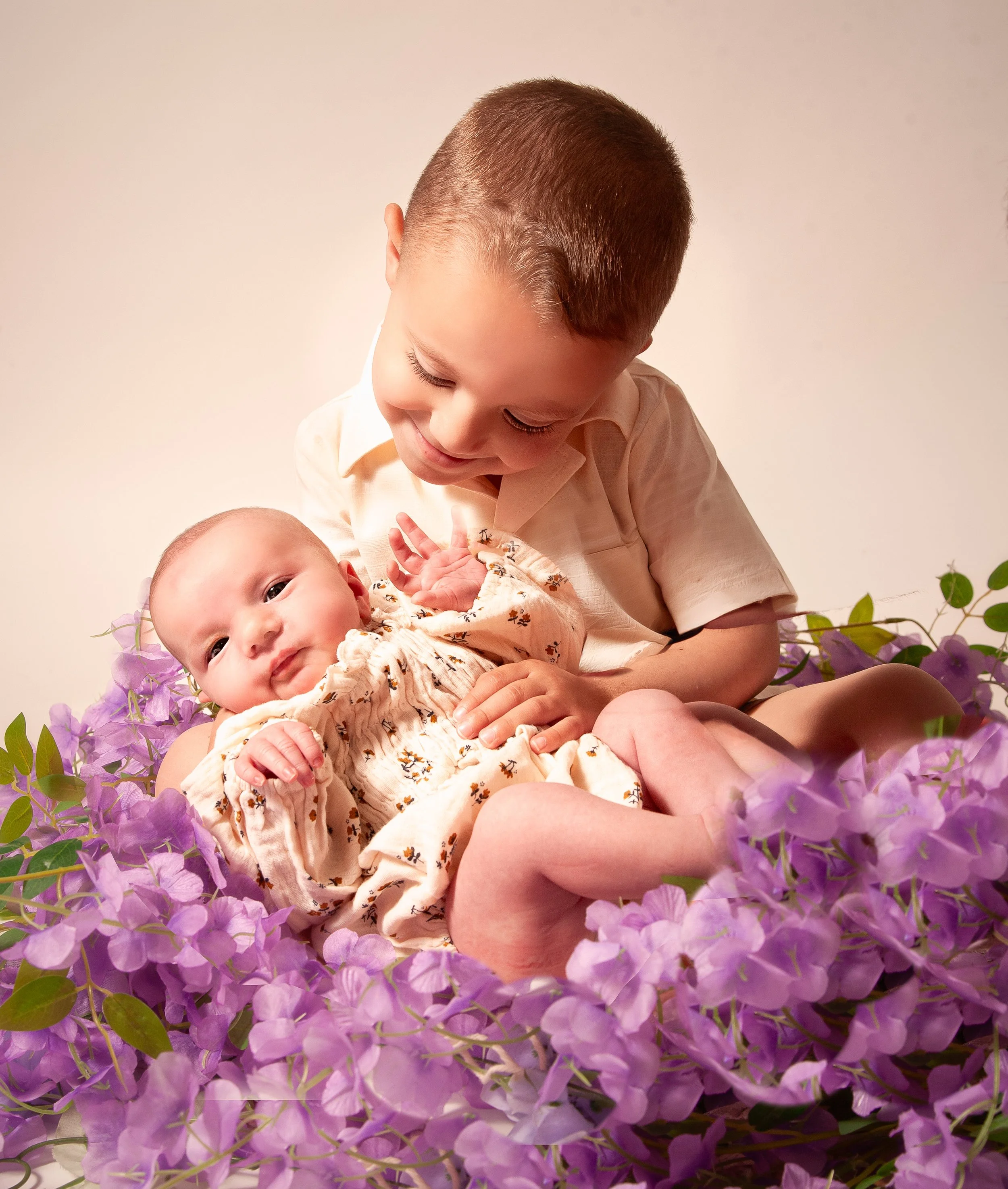 A young boy with a short haircut and a light-colored shirt smiling and looking down at a newborn baby he is holding. The baby is surrounded by purple flowers with green leaves, and has big eyes and a floral patterned outfit.
