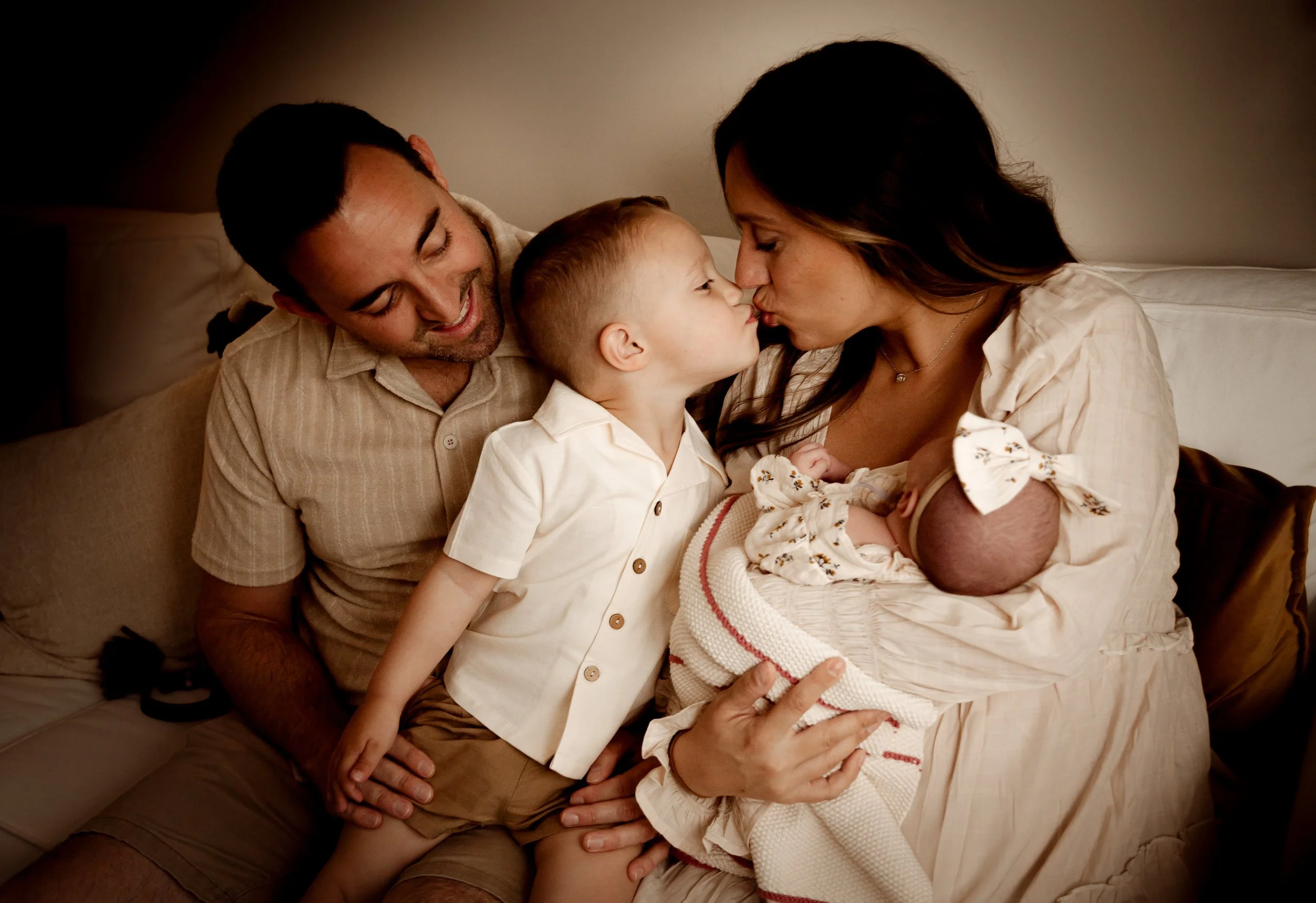 A family of three with a father, mother, and young son sitting on a sofa, leaning close and touching noses, sharing a tender moment.